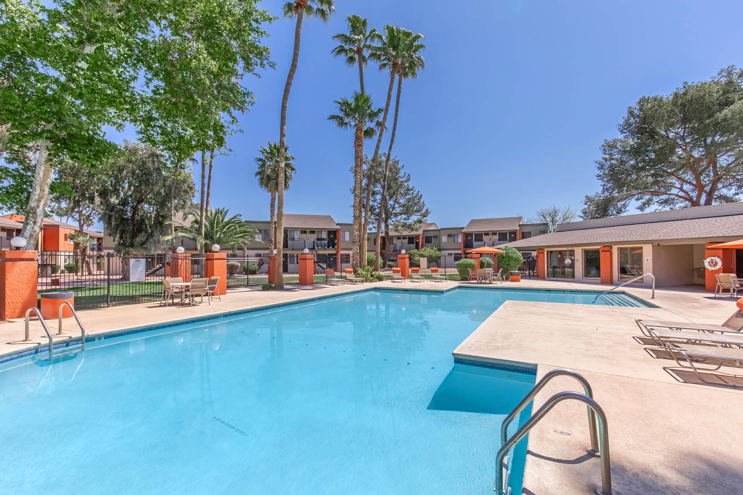 A bright outdoor swimming pool surrounded by palm trees and lush greenery. Lounge chairs and shaded umbrellas are positioned around the pool area. In the background, residential buildings can be seen, adding to the tranquil atmosphere of the setting.