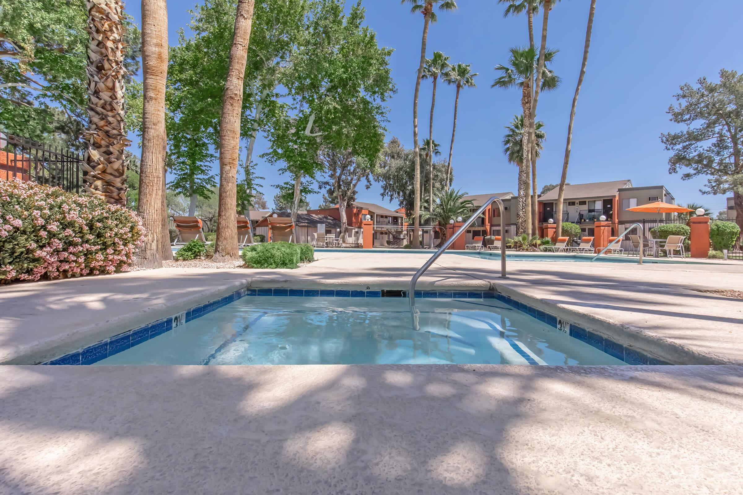 A clear hot tub surrounded by palm trees and lush greenery, with a bright blue sky overhead. In the background, there are lounge chairs and a vibrant pool area, creating a relaxing resort-like atmosphere.
