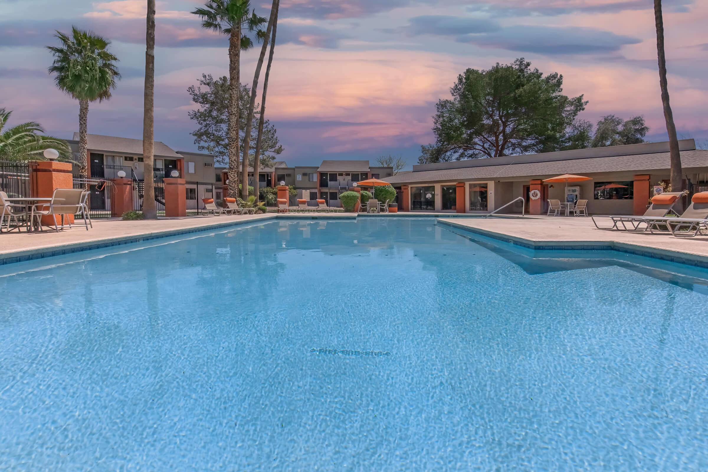 A tranquil outdoor swimming pool surrounded by palm trees and lounge chairs, reflecting a colorful sky at sunset. In the background, there are residential buildings with a modern design, creating a peaceful vacation atmosphere.