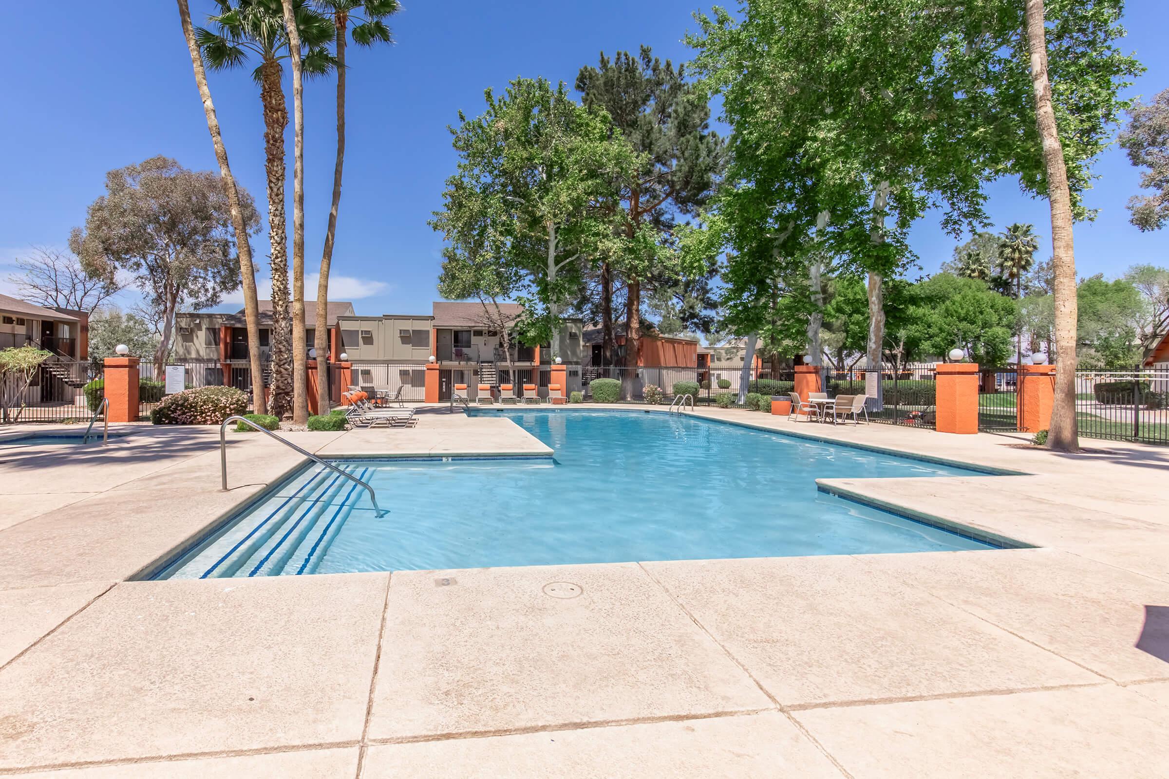 A clear blue swimming pool surrounded by a concrete patio, with lounge chairs nearby. Tall trees and green landscapes are visible in the background, along with residential buildings. The sky is bright blue with a few clouds, creating a sunny atmosphere ideal for relaxation.