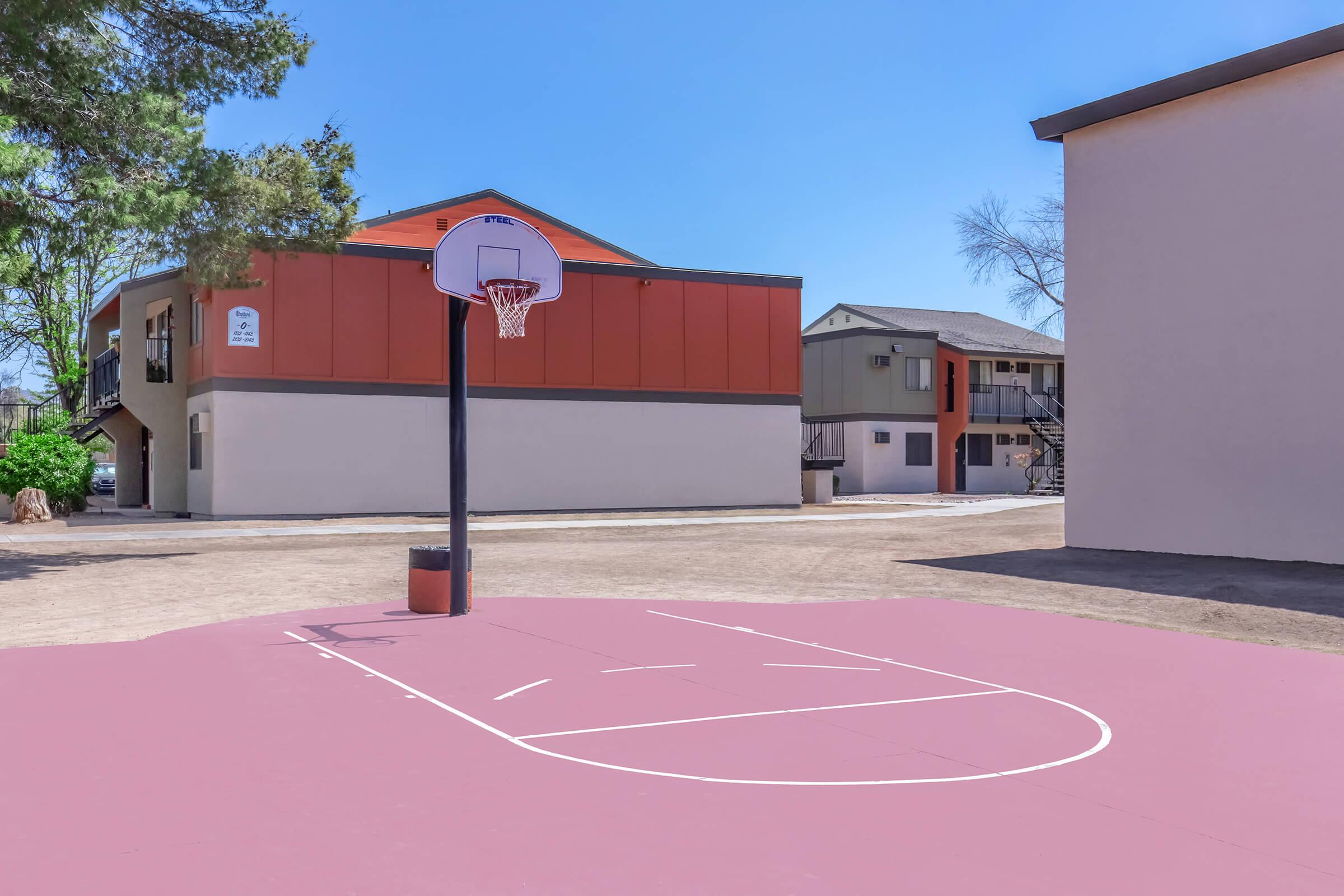 A basketball court with a hoop set against a backdrop of residential buildings. The court has a pink surface and is surrounded by dirt and grass. The sky is clear and blue, indicating a bright day.
