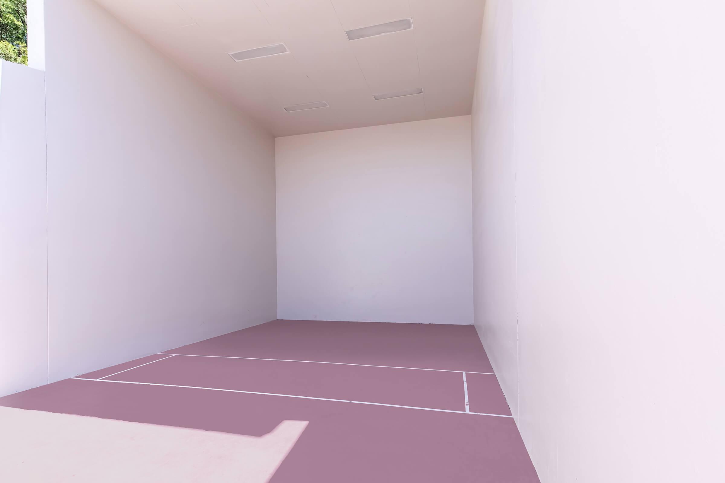 Empty indoor space featuring white walls and a pale pink floor. The room is well-lit with ceiling lights and appears to be a court or area designated for a sport, possibly squash or racquetball, given the markings on the floor.