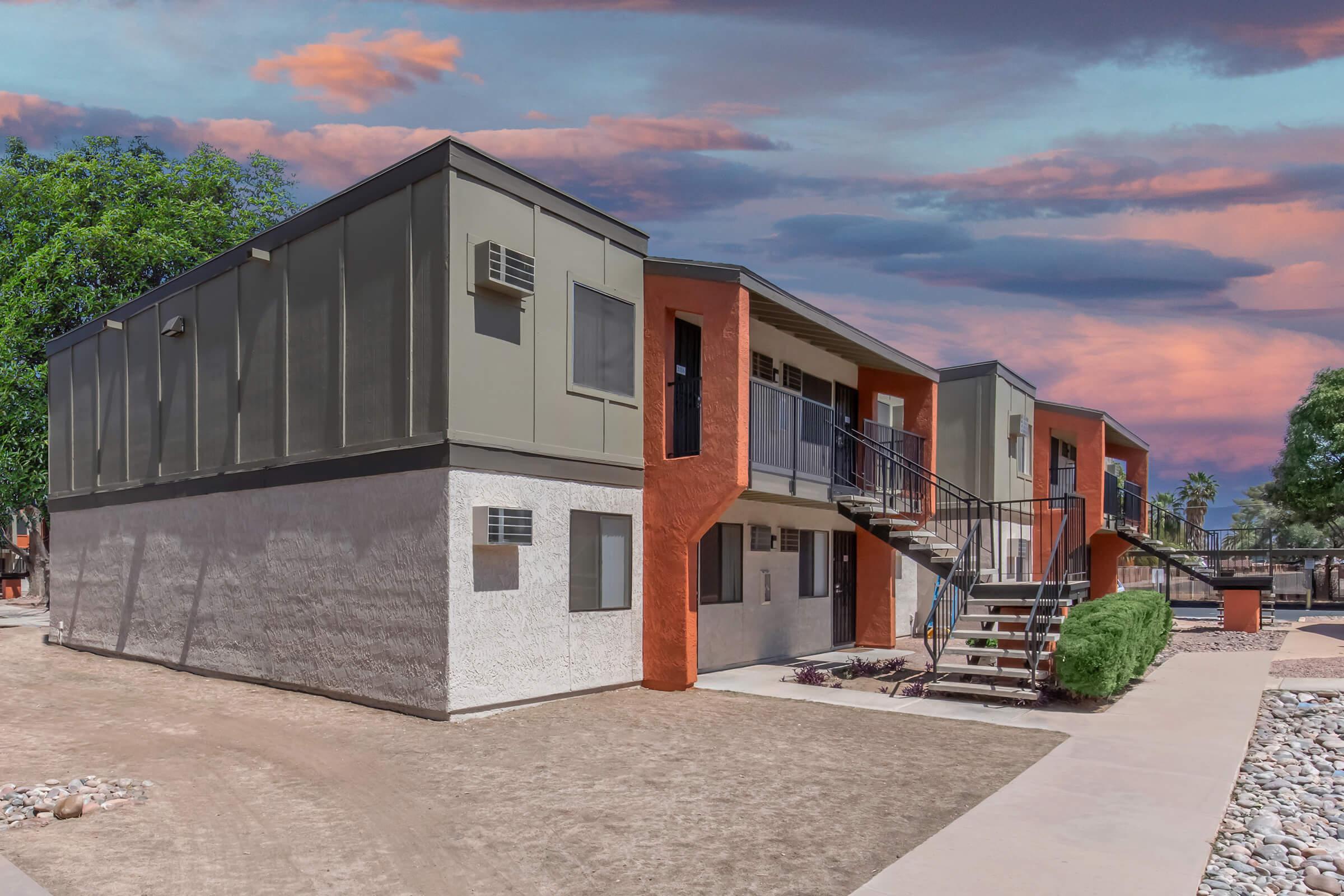Two-story apartment building with a modern design, featuring a mix of gray and orange colors. Each unit has a balcony and outdoor stairs. The surrounding landscape includes gravel paths, shrubs, and trees, with a colorful cloudy sky in the background.
