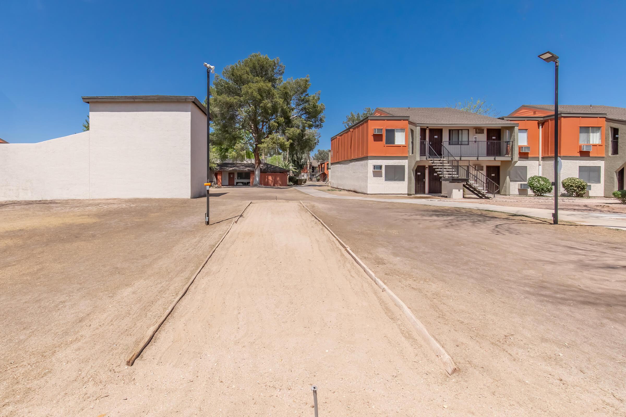 A wide view of a dusty pathway leading between two buildings in a residential area. On the left, there is a plain white wall, while on the right, two-story apartments are visible with orange and beige exteriors. The sky is clear blue, and a few trees and street lamps are present, creating an open, sunny atmosphere.