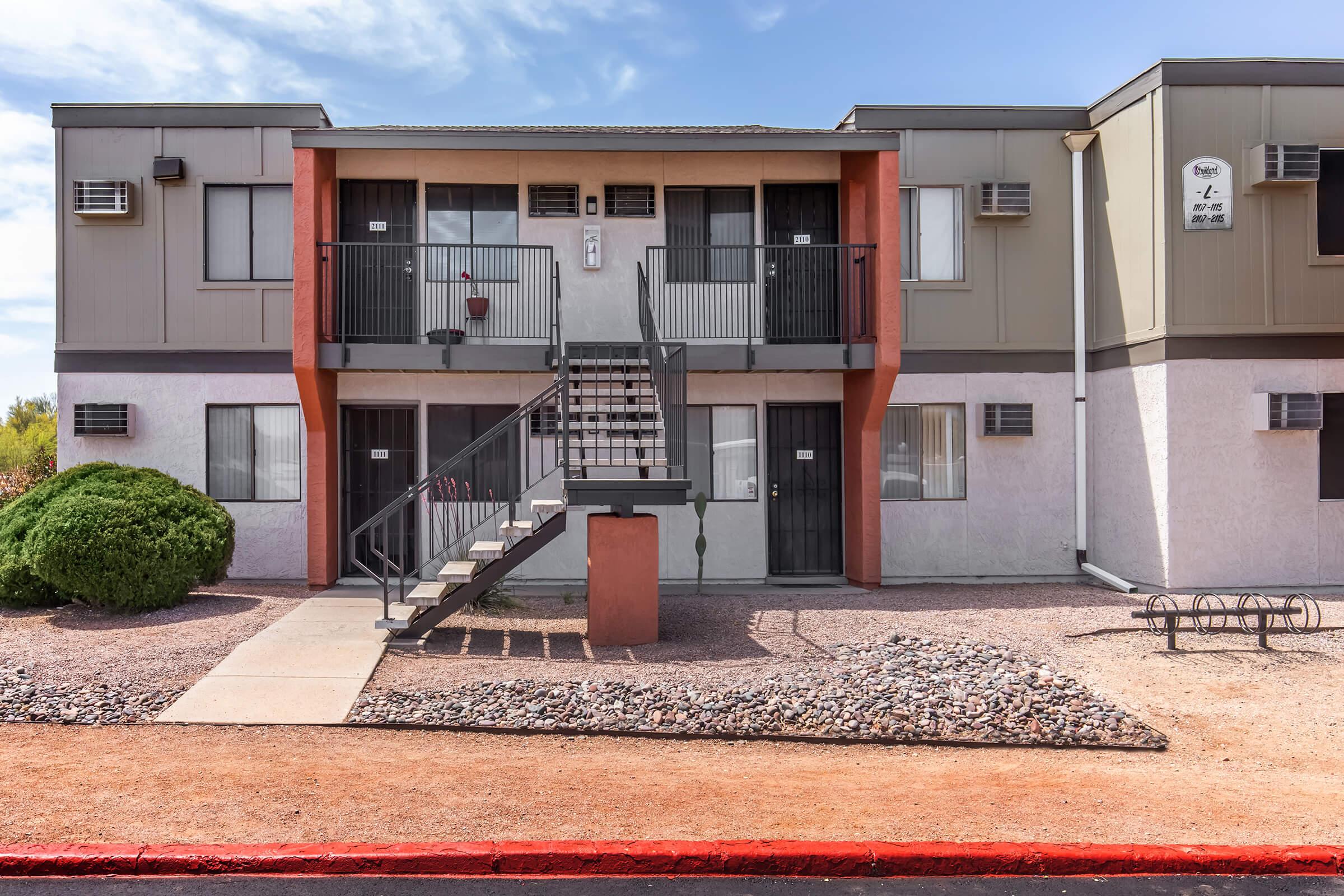 A two-story apartment building with a flat roof, featuring a central staircase leading to upper units. The façade includes a mix of beige, gray, and orange colors. Several air conditioning units are visible, and the landscape features low shrubs and gravel. There are bike racks positioned in front of the building.