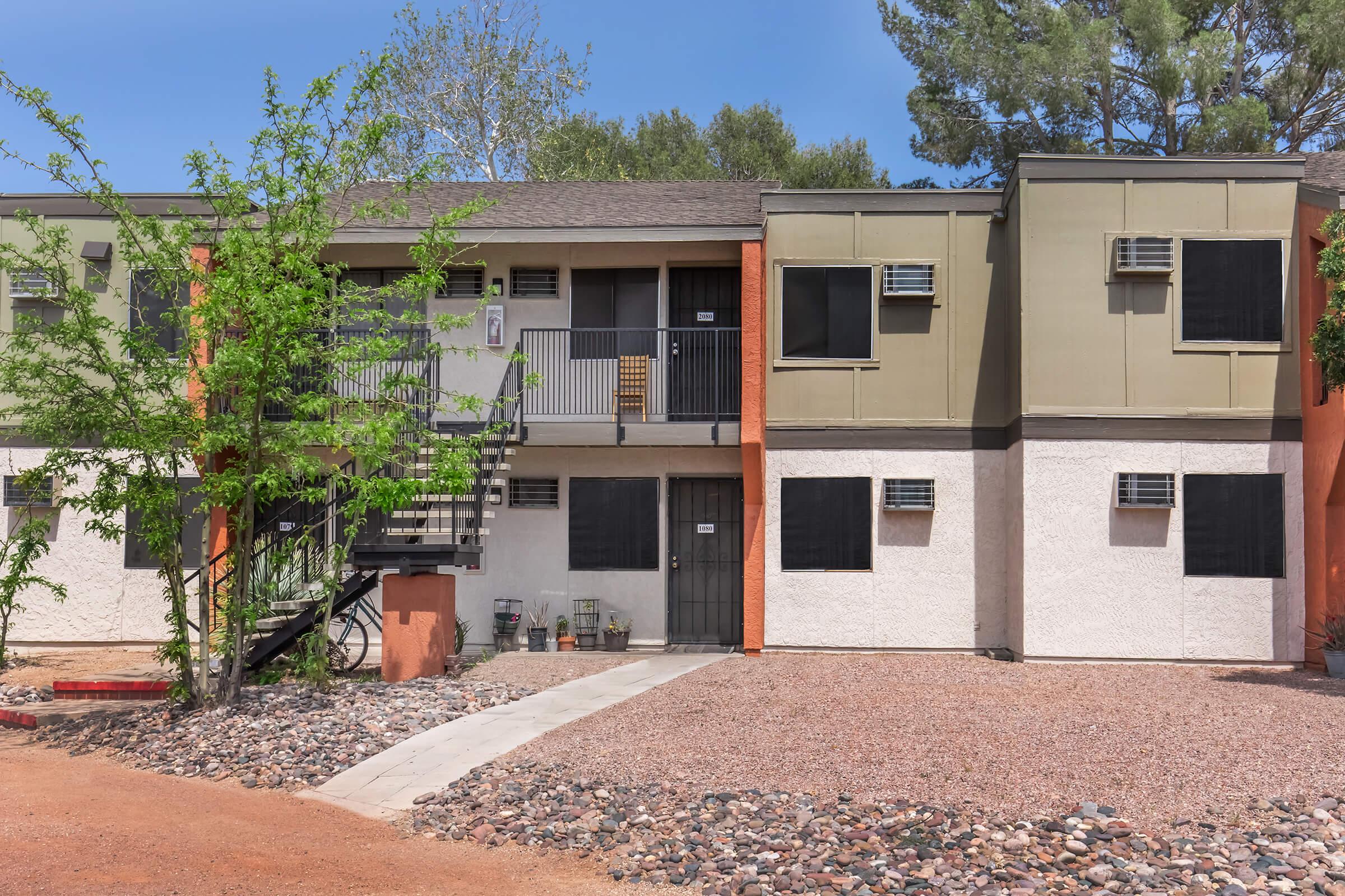 A two-story apartment building with sections in beige and orange. The building features external staircases, several air conditioning units, and blacked-out windows. Surrounding it are landscaped areas with small trees and gravel paths, set against a clear blue sky.