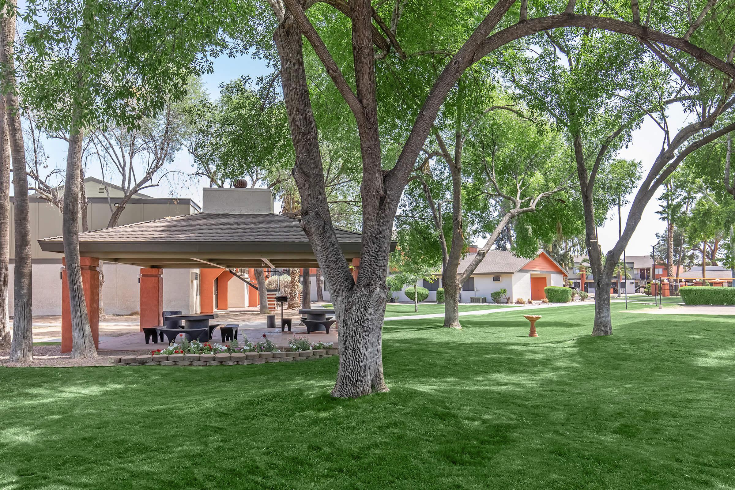 A well-maintained park featuring lush green grass, large trees providing shade, and a covered gazebo area. Picnic tables are visible beneath the gazebo, with manicured flower beds in the foreground. The background includes residential buildings and pathways, creating a serene outdoor environment.
