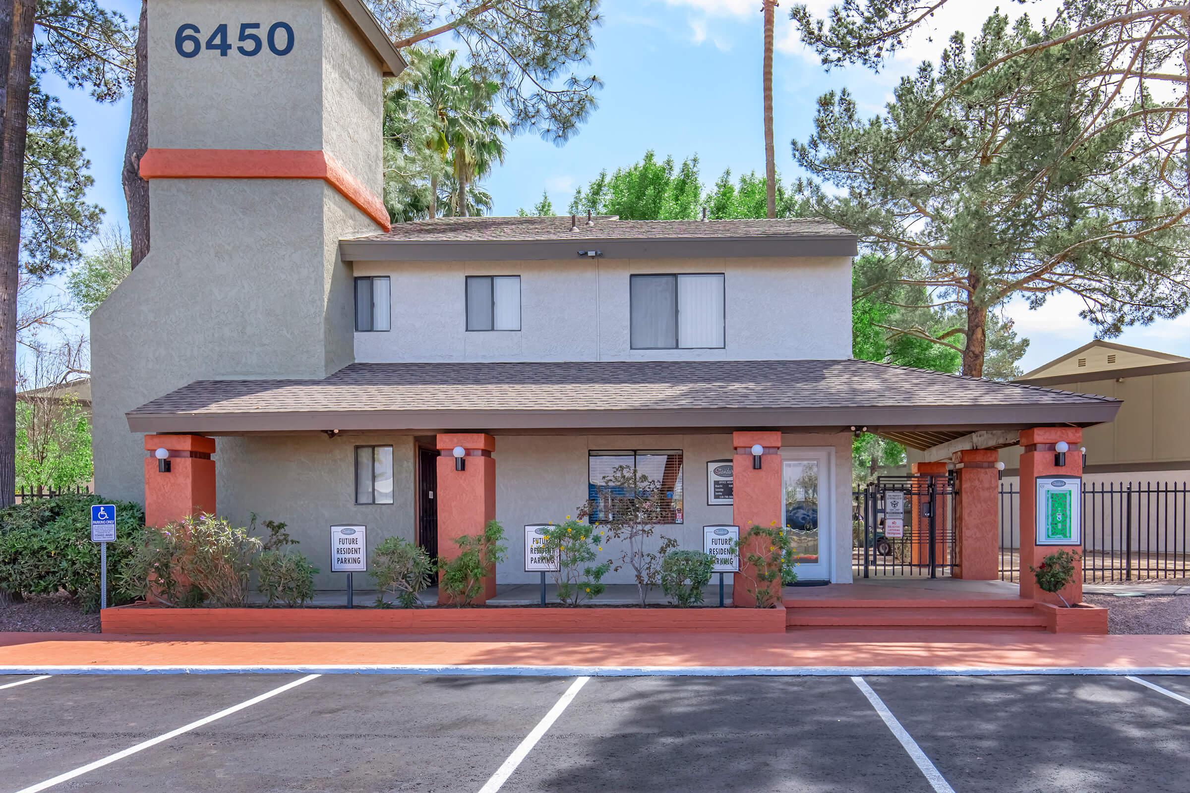 A two-story building with a sloped roof painted in light gray and orange accents. The entrance features a covered porch with pillars and a landscaped area. There are several parking spaces in front, and a sign displaying the building number "6450." Trees and greenery are visible in the background.