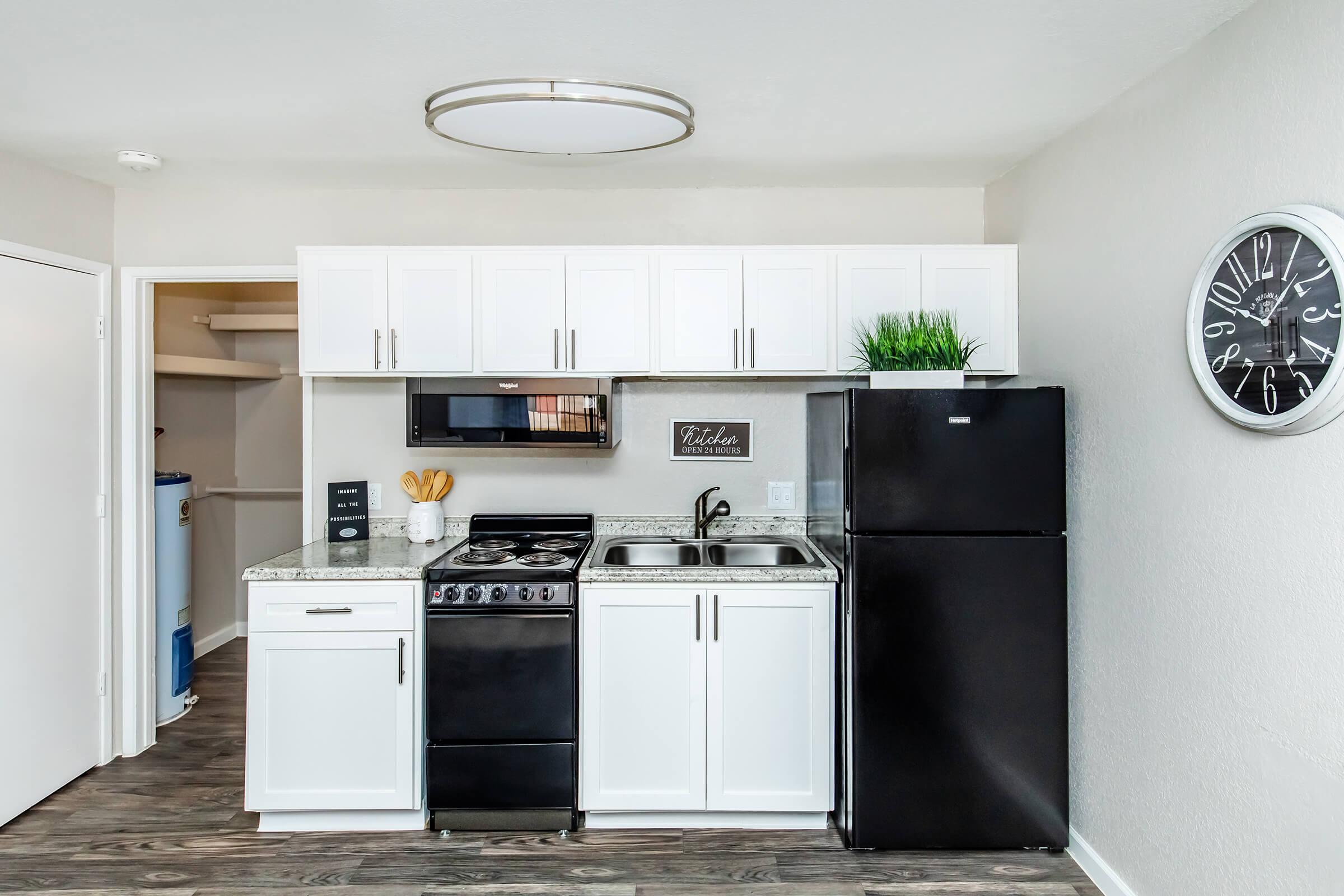 A modern kitchen featuring white cabinets, a black stove and refrigerator, a stainless steel microwave, and granite countertops. A clock on the wall and a plant decorate the space. A pantry door is visible in the background, enhancing the kitchen's functionality.