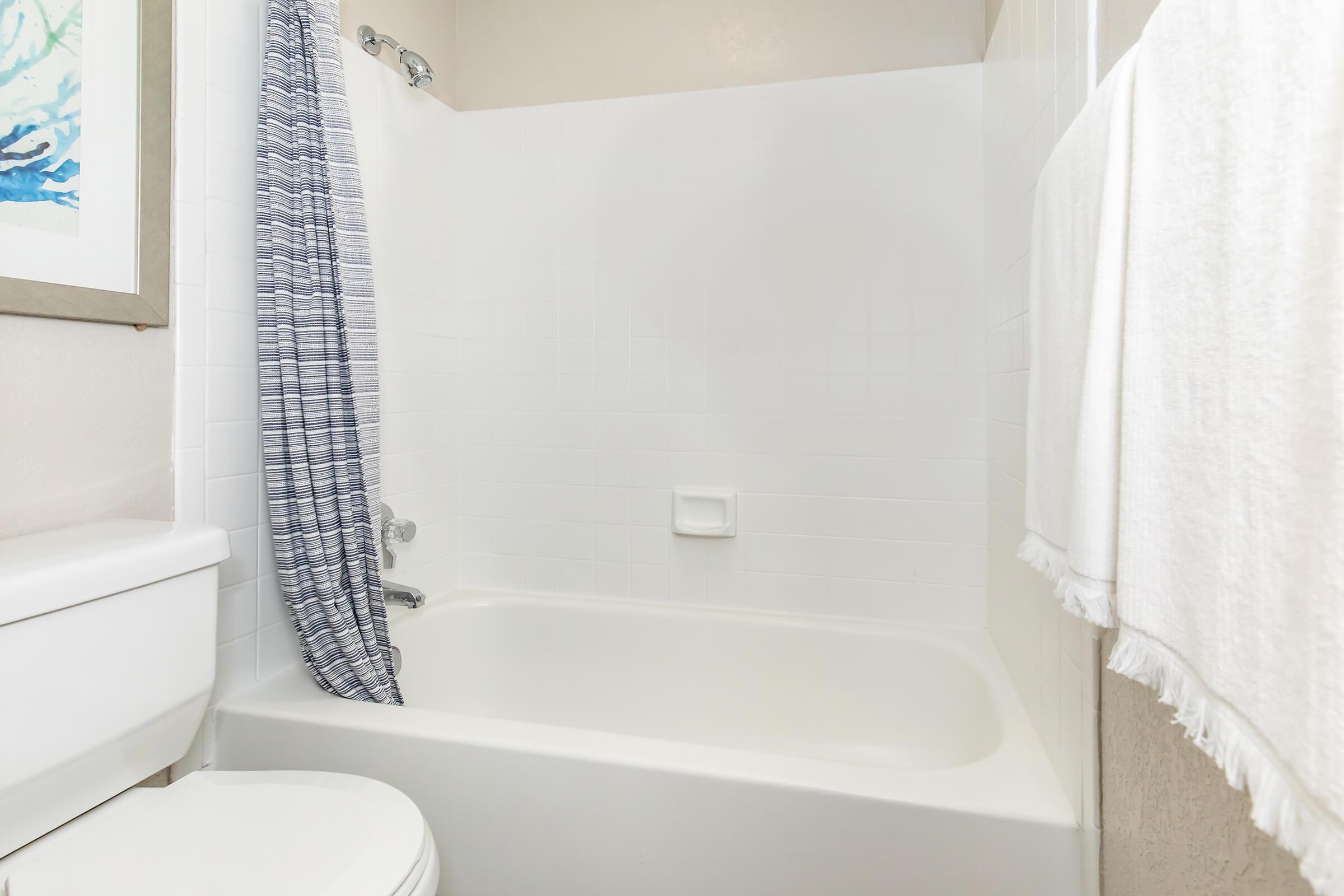 A clean, modern bathroom featuring a bathtub with a white shower curtain, a tiled wall, a white toilet, and a fluffy white towel hanging nearby. The decor is simple and neutral, contributing to a fresh, inviting atmosphere.