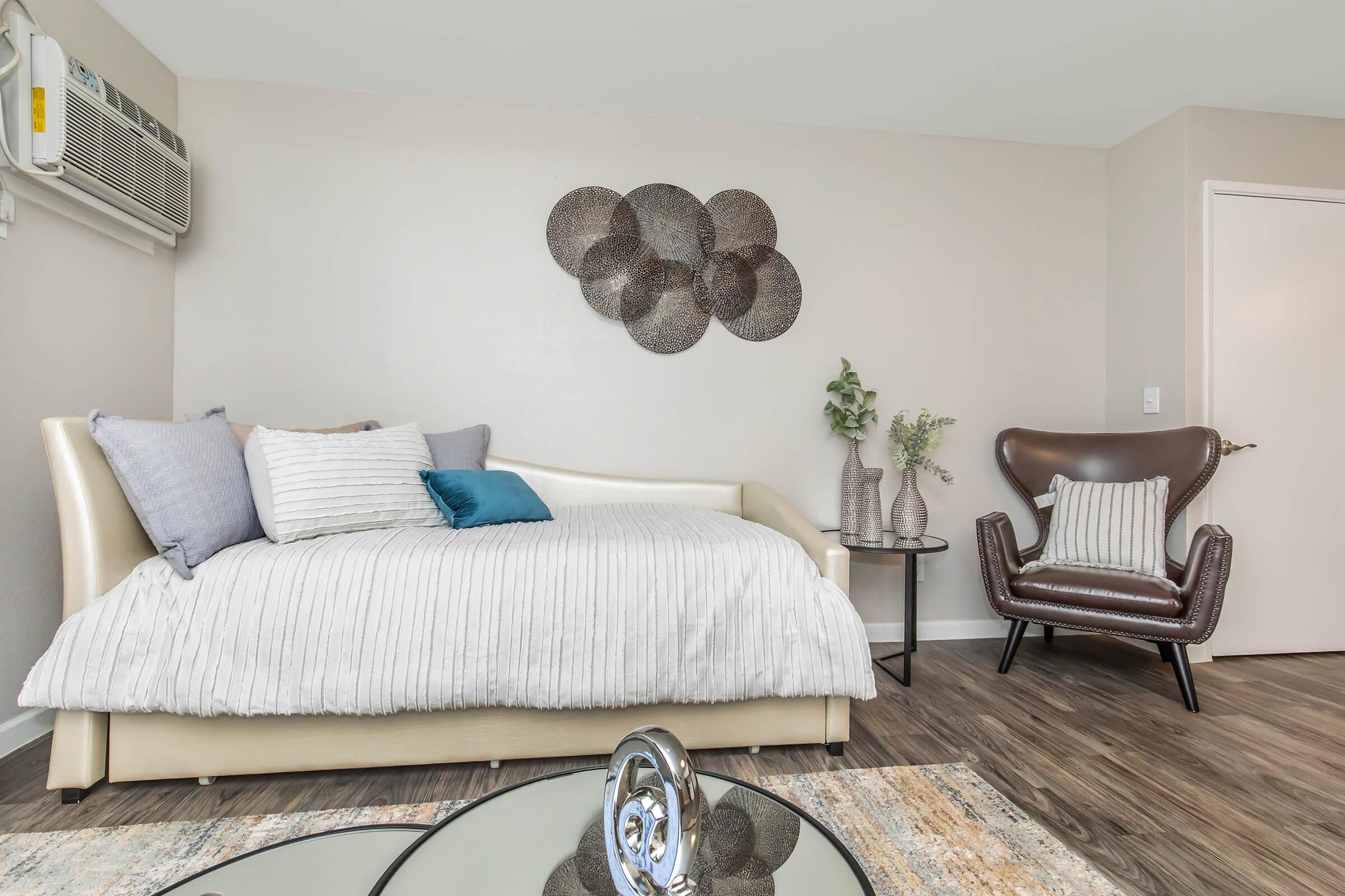 A cozy, modern living space featuring a light-colored daybed with gray and teal pillows, an elegant brown accent chair, and decorative wall art. There is a large round glass coffee table on a layered rug, with potted plants and vases enhancing the inviting atmosphere. A wall-mounted air conditioner is also visible.