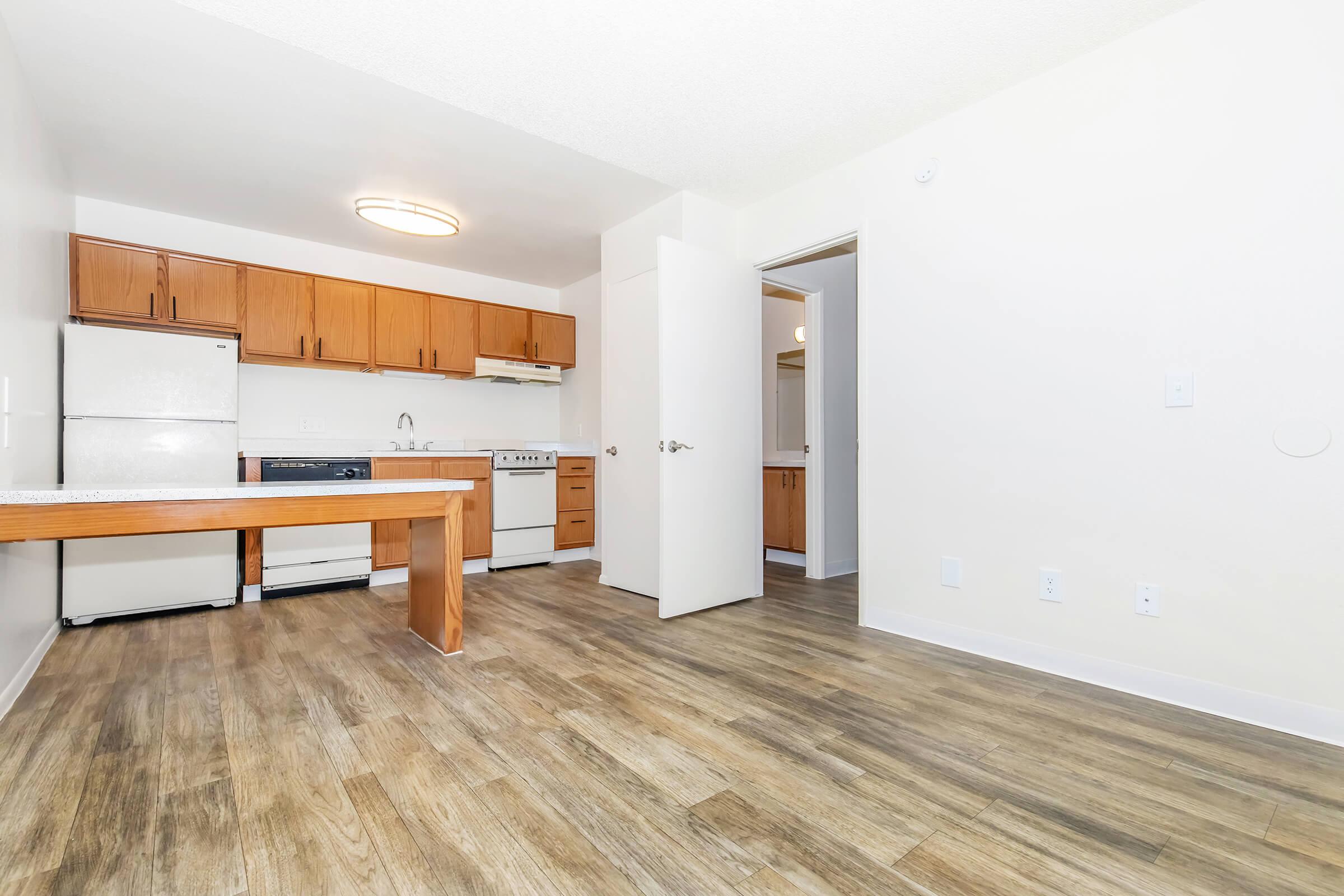 A modern kitchen and living area featuring wooden cabinets, a white refrigerator, and a stove. The space has laminate flooring and an overhead light fixture. A door leads to another room, and the layout is open, creating a bright and airy atmosphere.