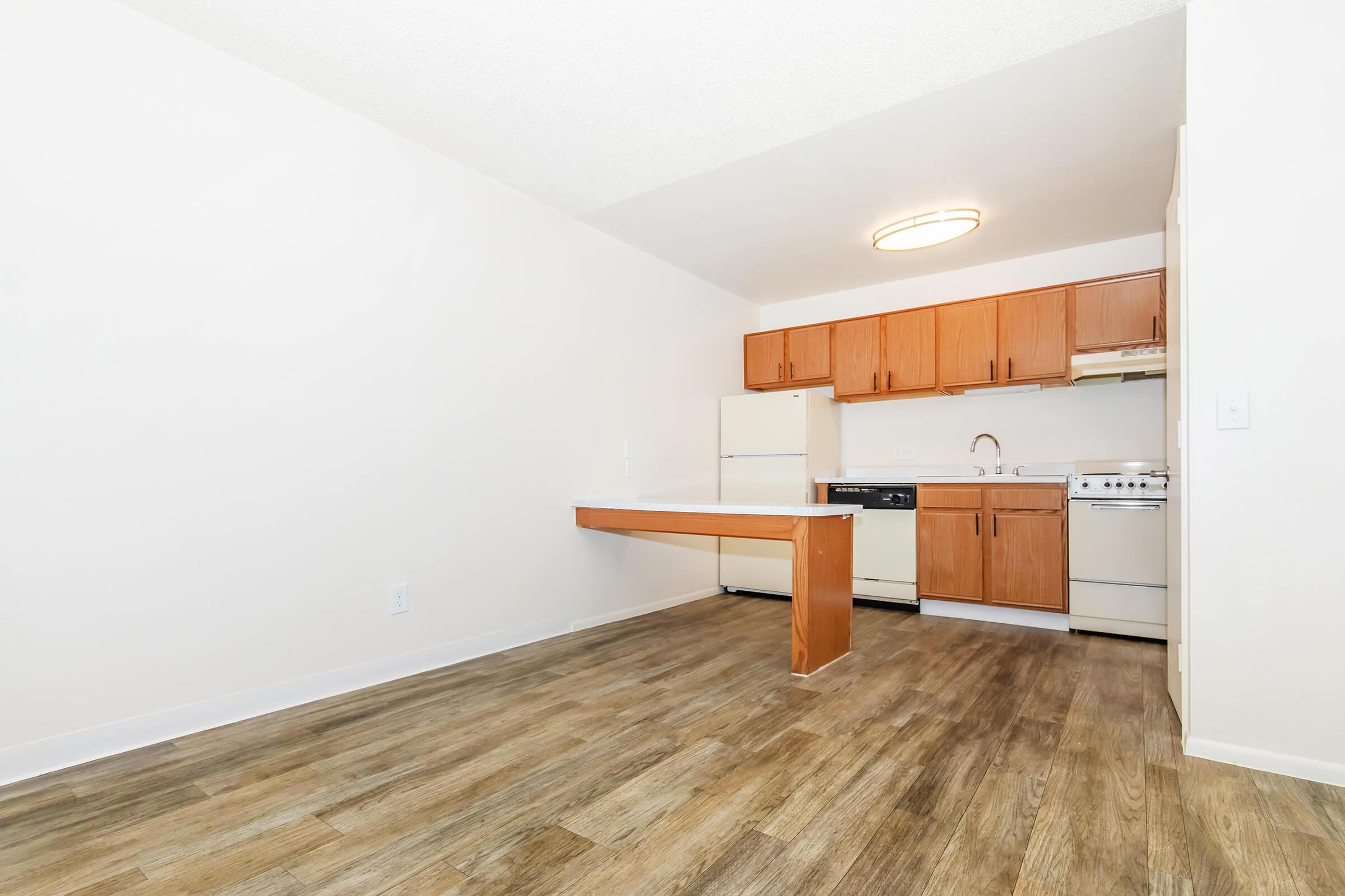 A modern and bright kitchen featuring wooden cabinets, a countertop with an overhang, and white appliances including a refrigerator, stove, and dishwasher. The flooring is a light wood finish, and the walls are painted white, creating an open and spacious feel. Natural light illuminates the area from above.