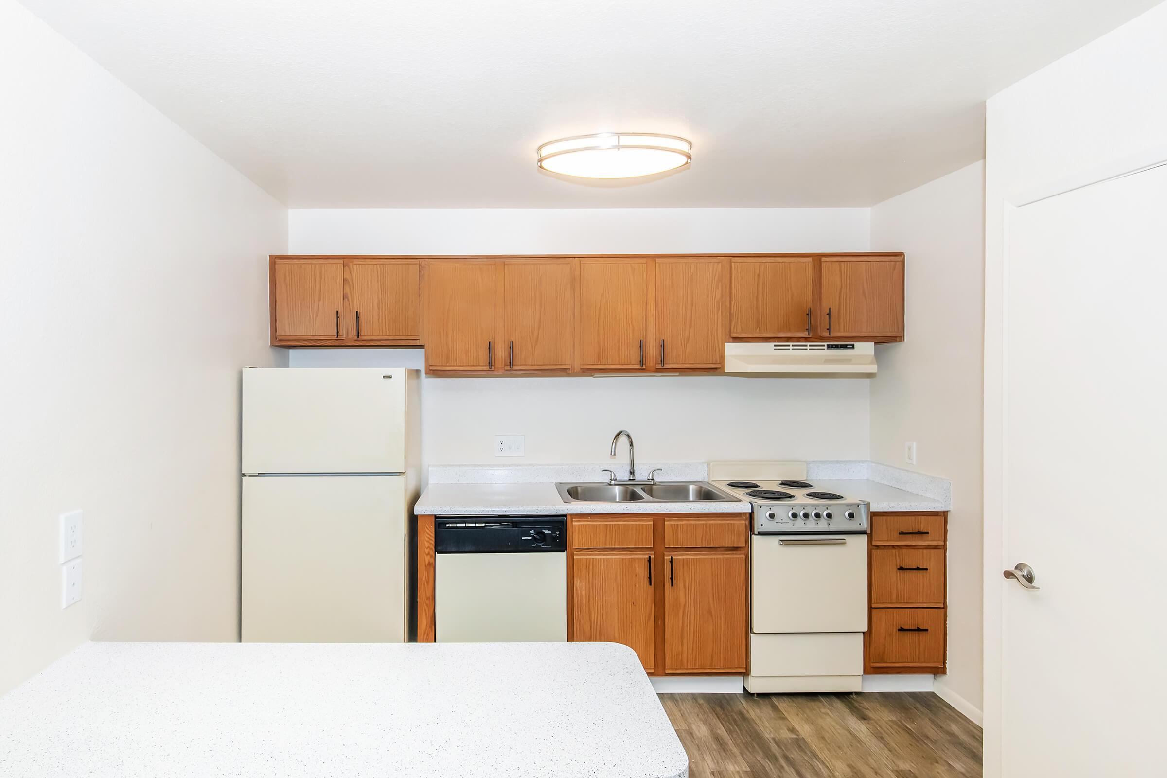 Modern kitchen featuring wooden cabinets, a refrigerator, dishwasher, and stove. The countertop is light-colored, complementing the white walls. A ceiling light fixture illuminates the space, highlighting the clean and functional layout. laminate flooring adds warmth to the area.