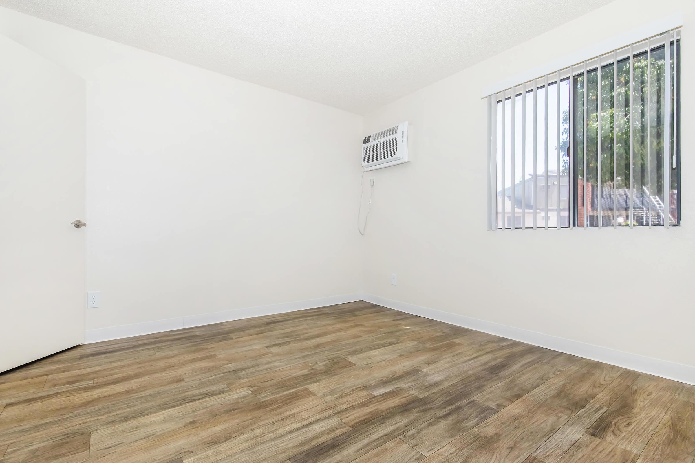 A vacant room with light-colored walls and a wooden floor. A window with vertical blinds allows natural light in, and there is a wall-mounted air conditioning unit. The door is partially visible on the left side of the image. The overall space appears clean and unfurnished.