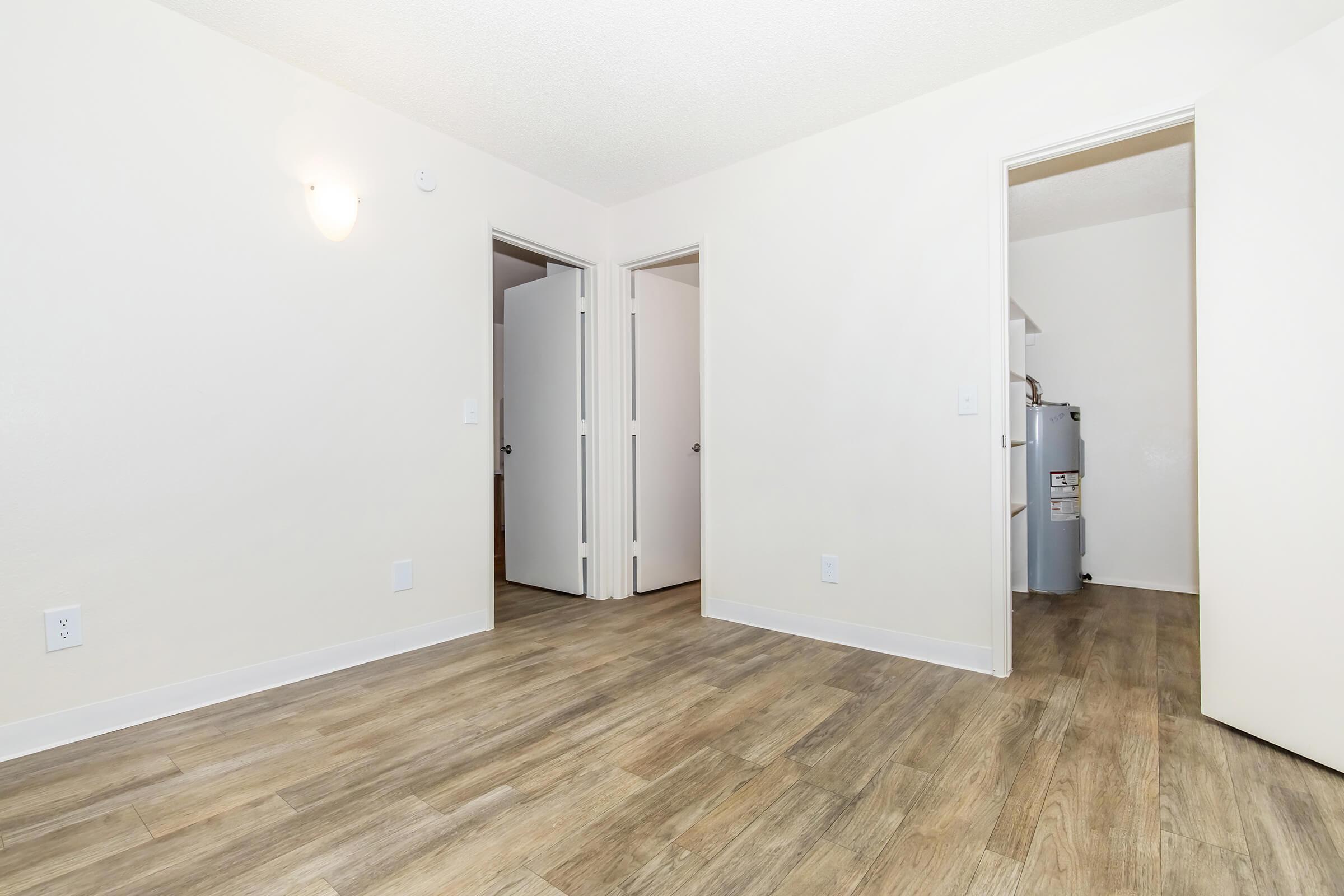 Empty room with light-colored walls and wooden flooring. There are two closed doors leading to different spaces, and a small door visible in the background. Soft lighting illuminates the area, creating a spacious and neutral environment.