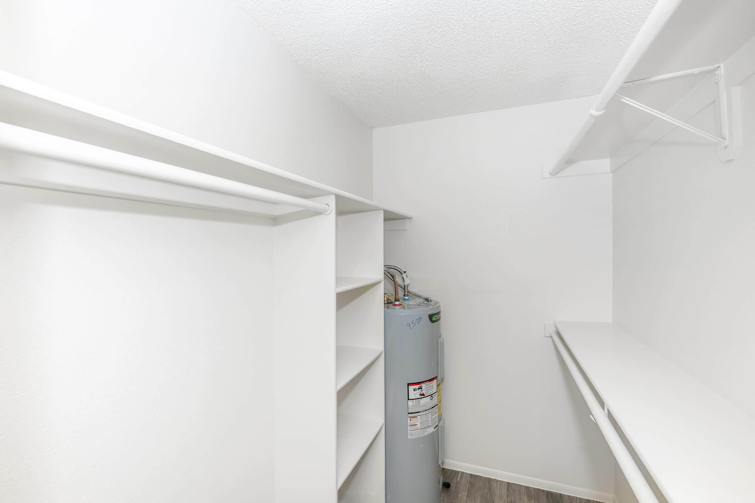 A clean, empty closet space featuring white walls, shelves on either side, and a water heater against one wall. The floor is a light wood-like material, and there is ample natural light illuminating the area. The overall design is simple and functional, ideal for storage.