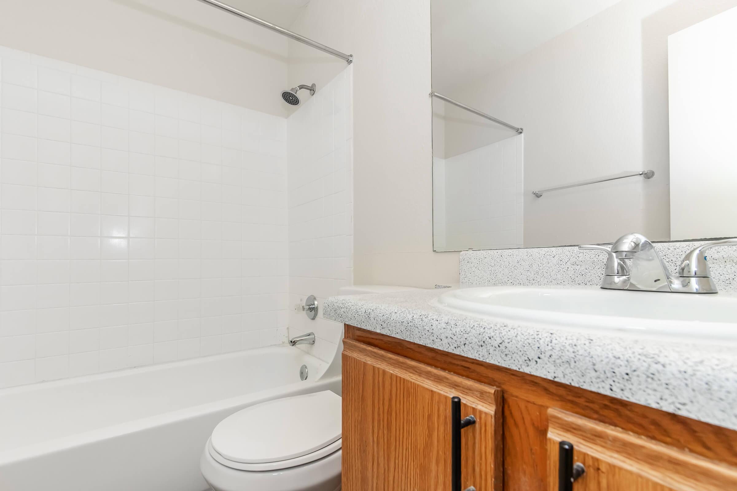 A clean, modern bathroom featuring a white tiled shower and bathtub, a wooden cabinet with sink, and a mirror. The walls are painted in a light color, enhancing the spacious feel. A towel bar is mounted on one wall, adding functional storage.