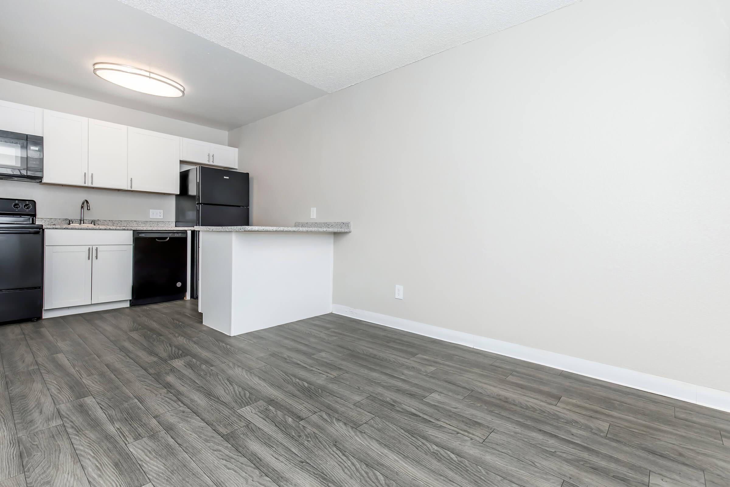 A modern kitchen featuring white cabinets, a black refrigerator, and a gray countertop. The space has light-colored walls and wood-style flooring, creating a bright and inviting atmosphere. A circular ceiling light provides illumination, enhancing the clean and minimalist design.