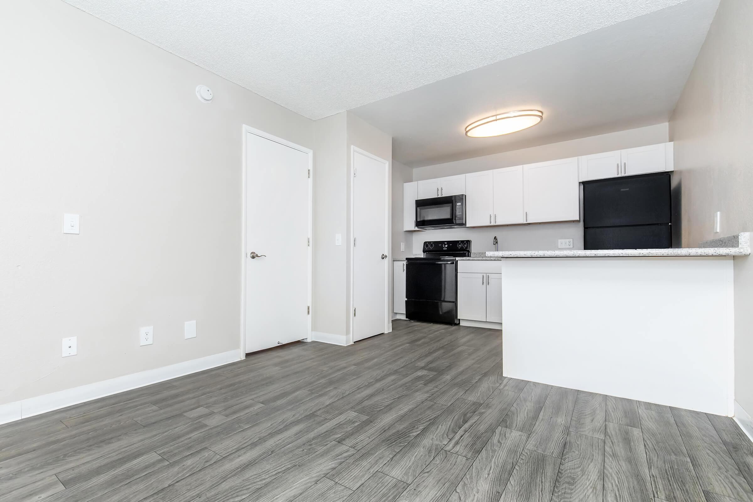 Modern kitchen and living area in a well-lit apartment. Features light gray walls, wood-style flooring, a black stove and refrigerator, and white cabinets. A clean, open layout with a central island and ample natural light from a ceiling fixture.