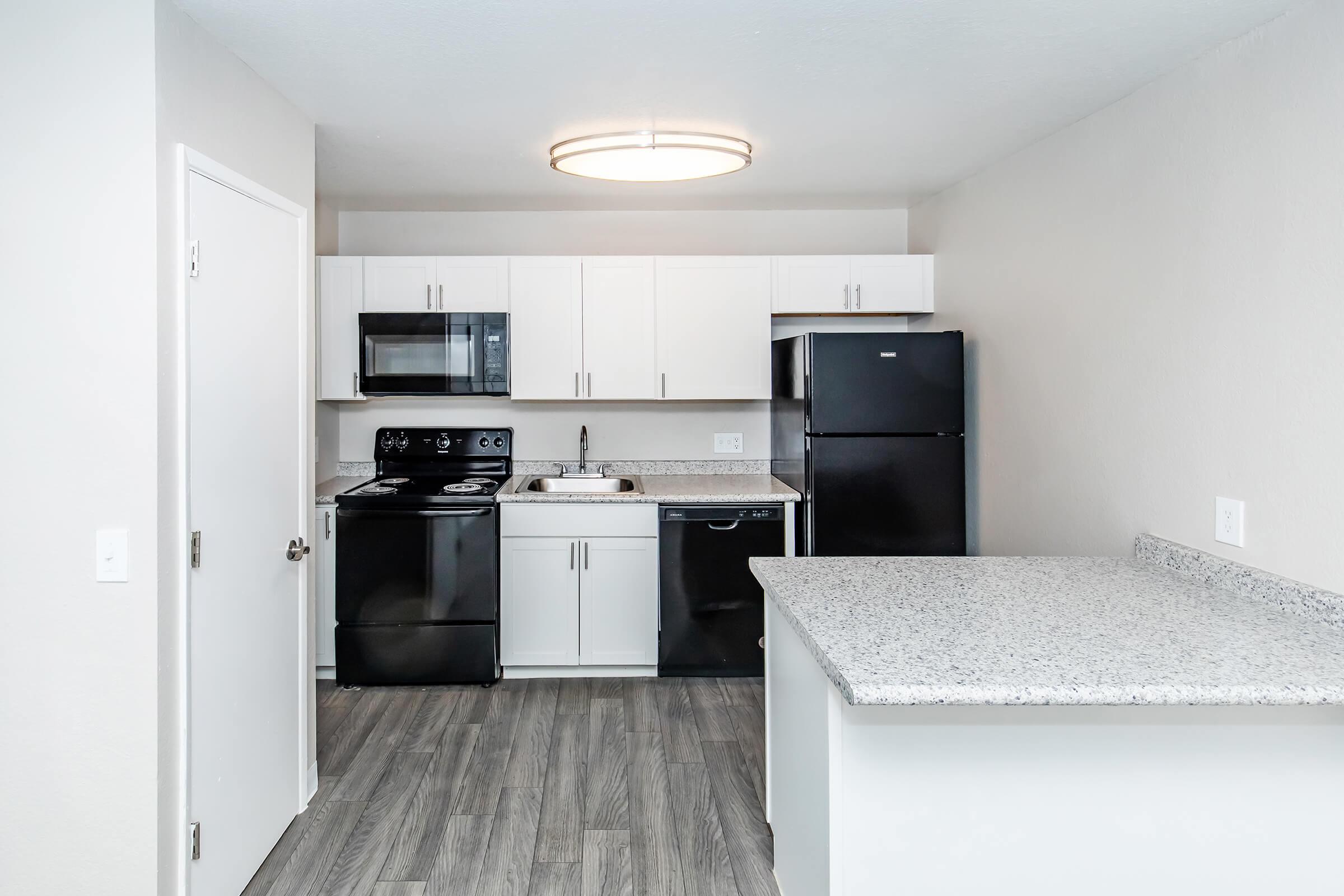 A modern kitchen featuring black appliances, including a stove and refrigerator, along with white cabinets and a granite countertop. The space has a light-colored wall and laminate flooring, illuminated by a circular ceiling light. The layout appears open and functional, suitable for cooking and dining.