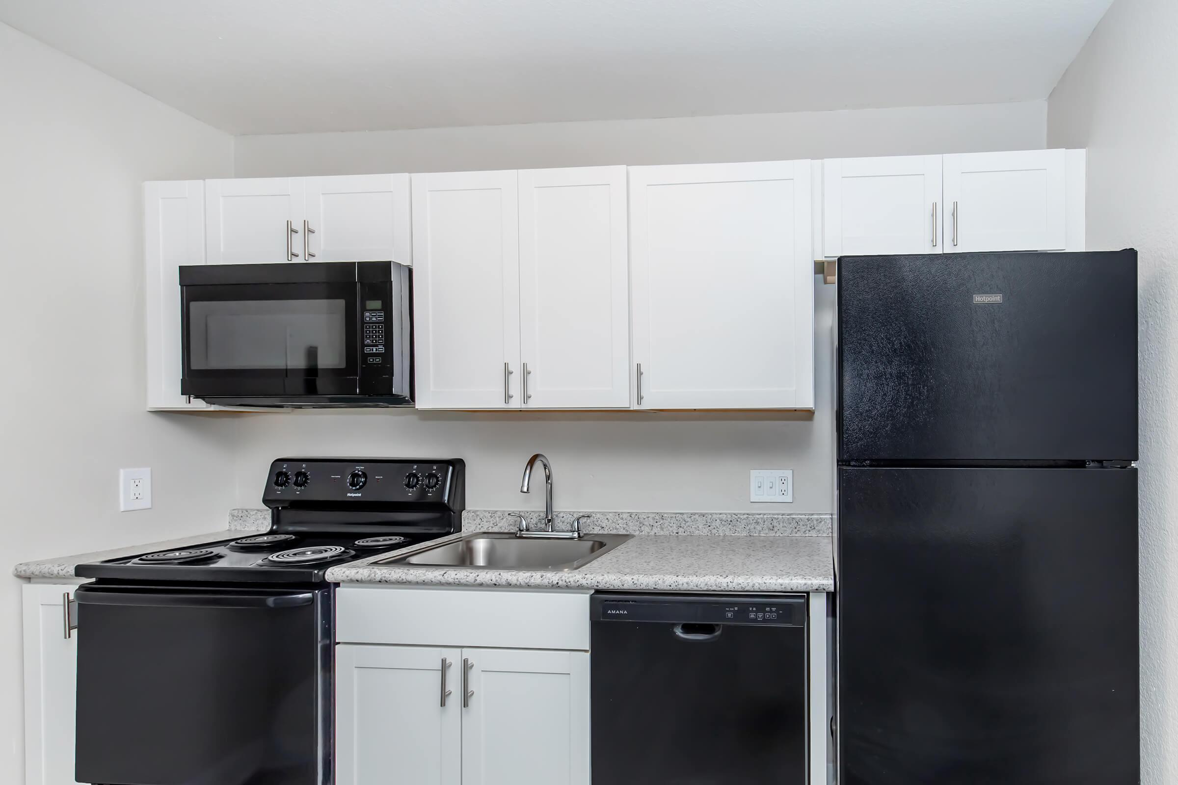 A modern kitchen featuring white cabinets, granite countertop, and black appliances. It includes a refrigerator, microwave, stove, and dishwasher. The kitchen has a clean and organized appearance, with a single sink and ample cabinet space for storage.