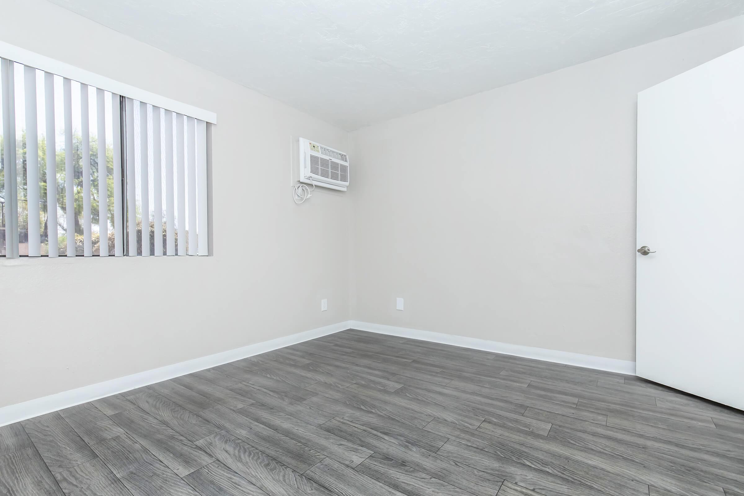 A light-colored room with gray wood flooring, featuring a partially open white door and a window with vertical blinds. An air conditioning unit is mounted on the wall beside the window. The walls and ceiling are painted in neutral tones, creating a clean and open space.