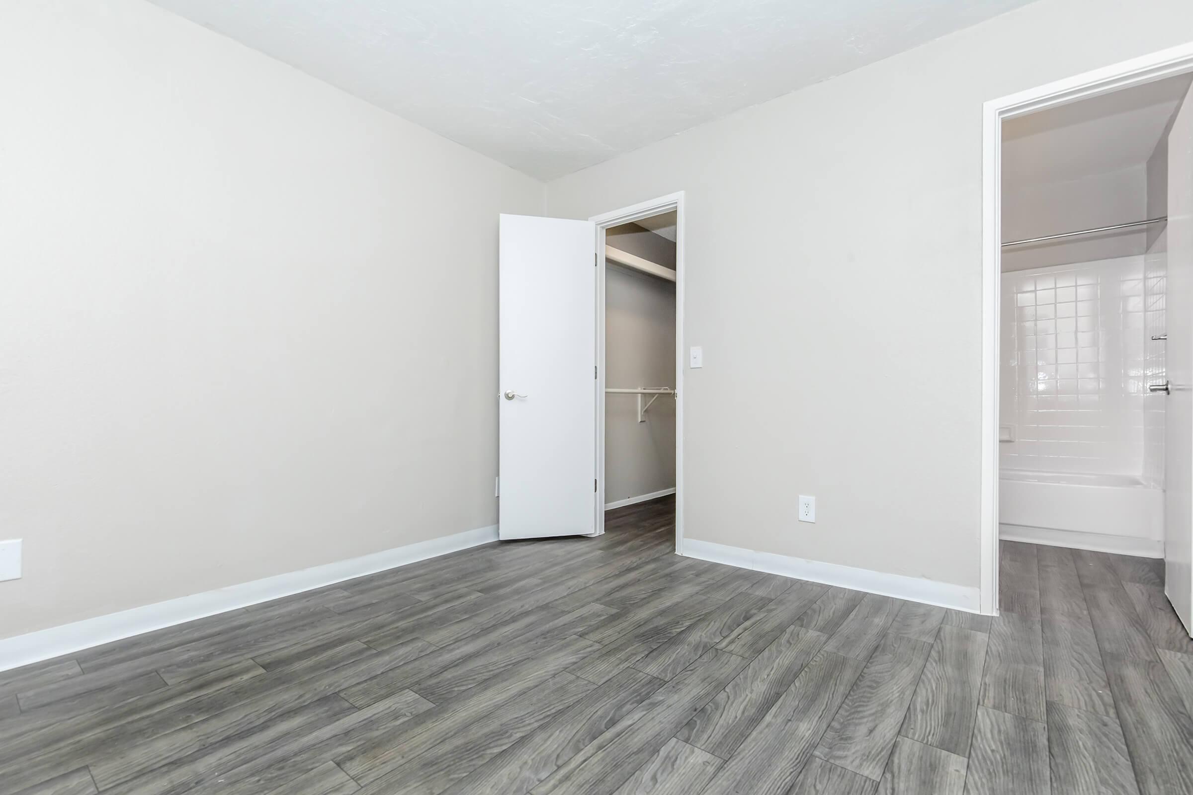 Interior view of a vacant room with light gray walls and laminate flooring. The room features an open doorway leading to a closet and another doorway that leads to a bathroom with white tiles. Natural light fills the space, giving it a bright and airy feel.