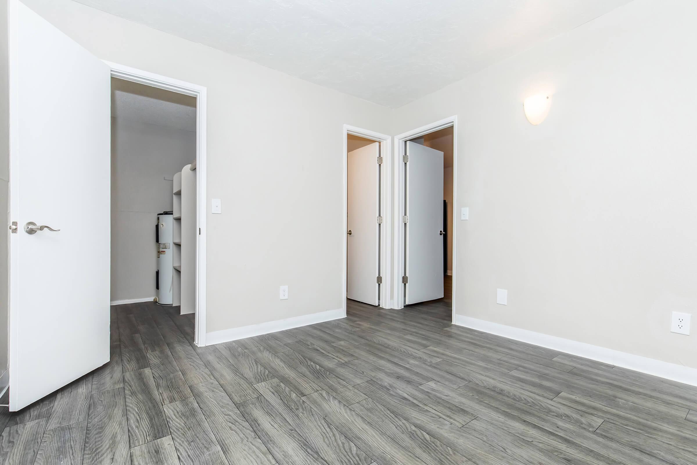 A light-filled room with gray wood laminate flooring. Two open doors lead to adjoining spaces, while a wall-mounted light fixture provides illumination. The walls are painted in a neutral color, creating a clean and inviting atmosphere. A small glimpse of plumbing equipment is visible in one doorway.