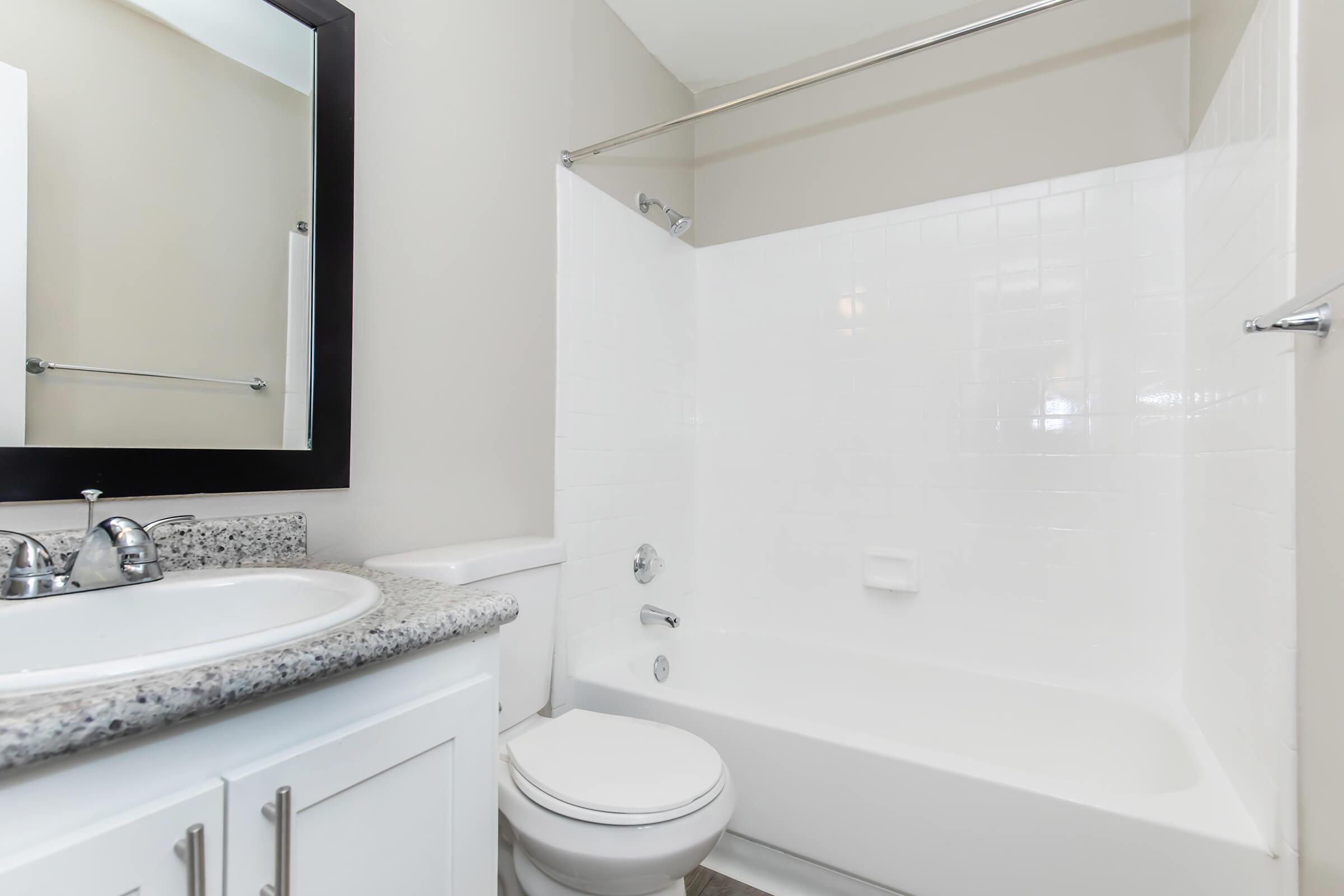 A clean, bright bathroom featuring a white bathtub and shower combination. The countertop is made of gray granite, and there is a round mirror above the sink. The walls are painted in a light color, creating a spacious feel. A towel rack is mounted on the wall, and the floor is tiled.