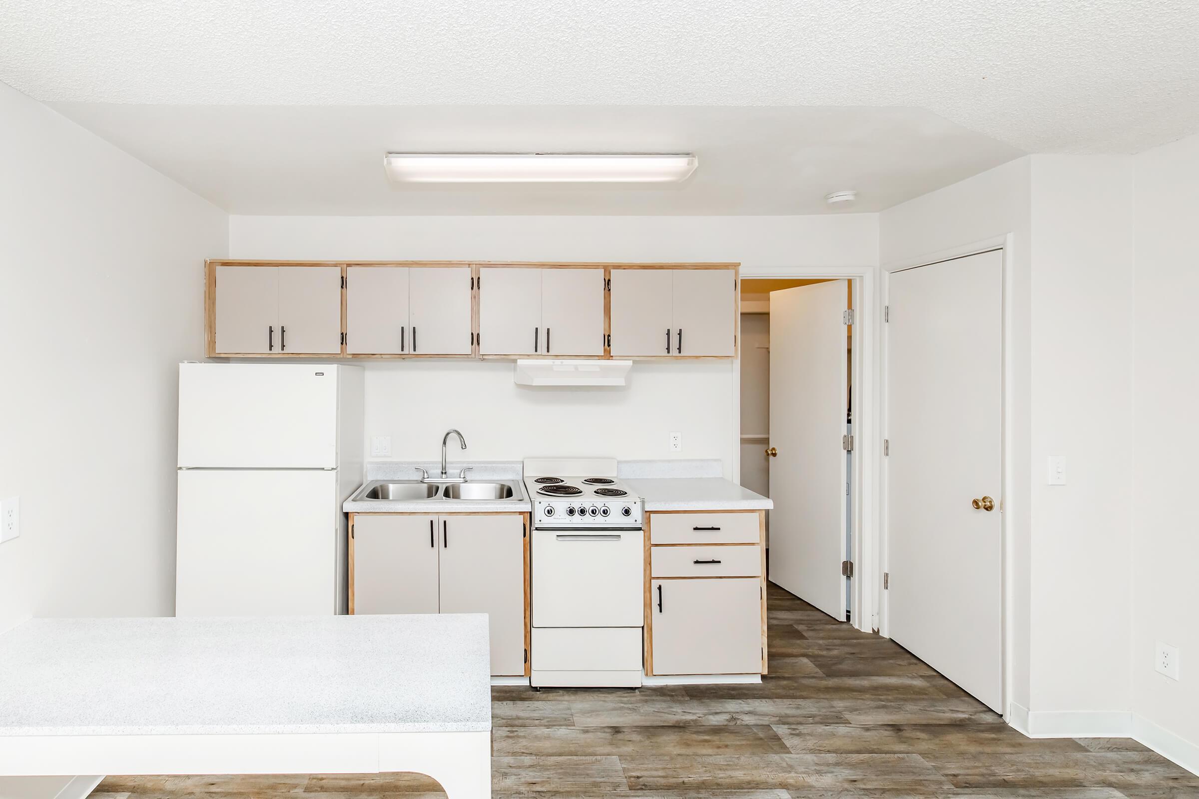A clean and modern kitchen featuring a white refrigerator, a sink, a stove, and cabinets. The countertops are light-colored, and the flooring is wood-style. The space is well-lit with a ceiling light, and a door leads to another room.