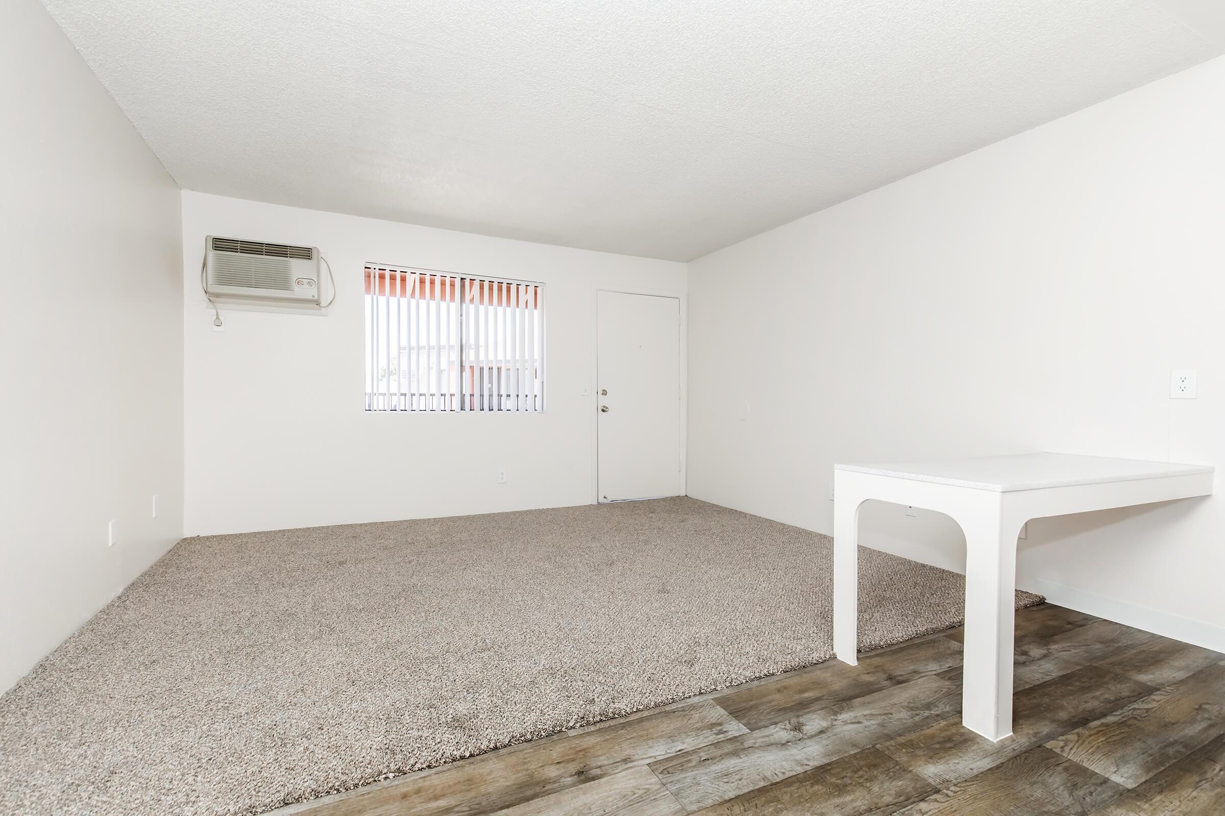 An empty room featuring light-colored walls, a window with blinds, and a door. There is a small table in one corner, while the floor is covered with beige carpeting and wooden planks. An air conditioning unit is mounted on the wall near the window.