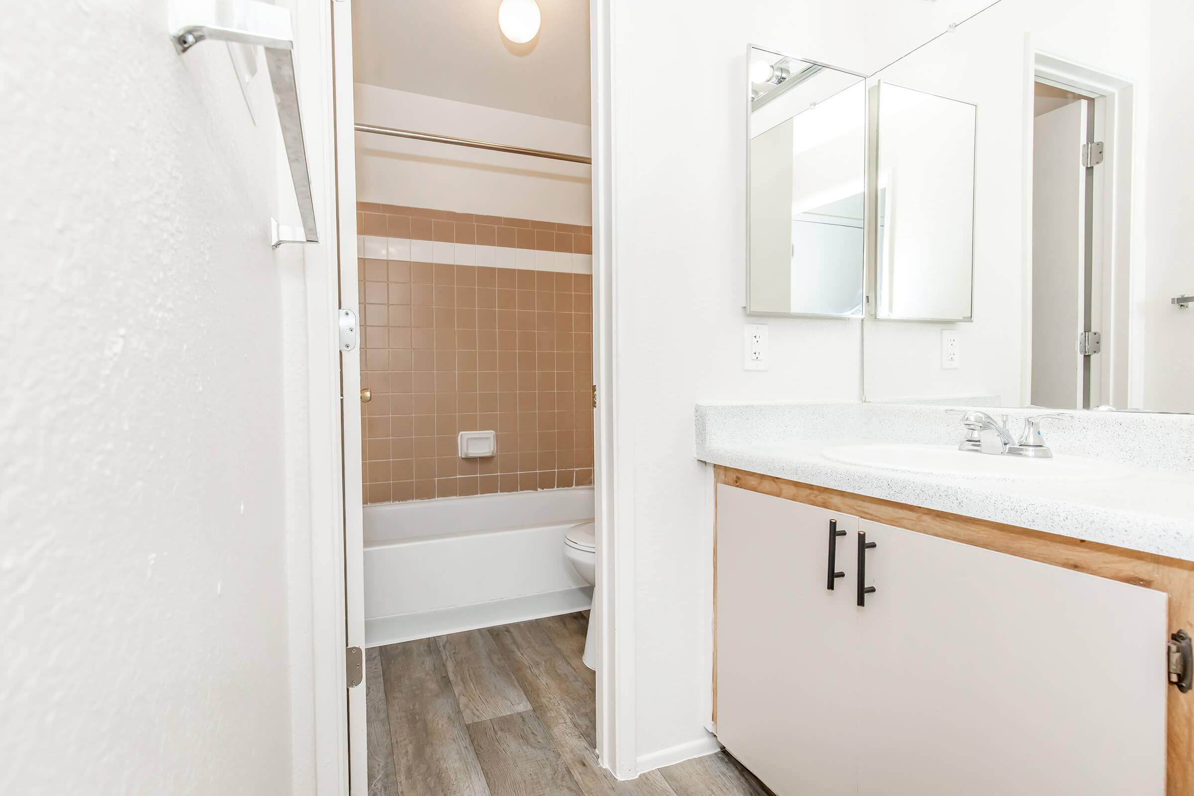 A clean, modern bathroom featuring a light-colored wall, a spacious vanity with a countertop, two mirrors above the sink, and a bathtub with a tiled backsplash. The flooring is laminate, and there’s a shower curtain rod visible. Natural light comes from a ceiling fixture.