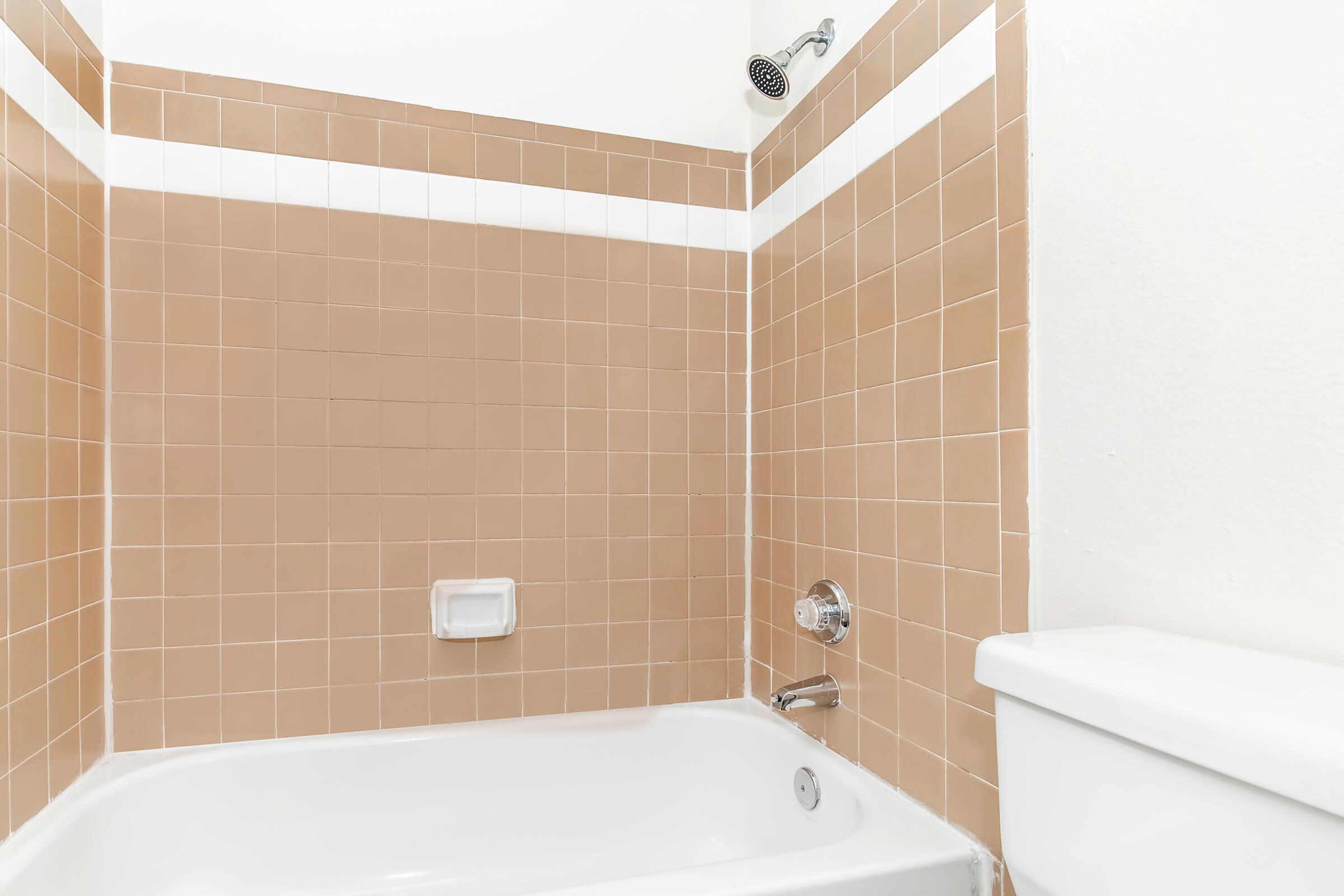 Clean bathroom with a bathtub and showerhead. The walls are tiled with brown and white ceramic tiles. A white toilet is visible on the right side of the image, and the area is well-lit, creating a tidy and simple aesthetic.