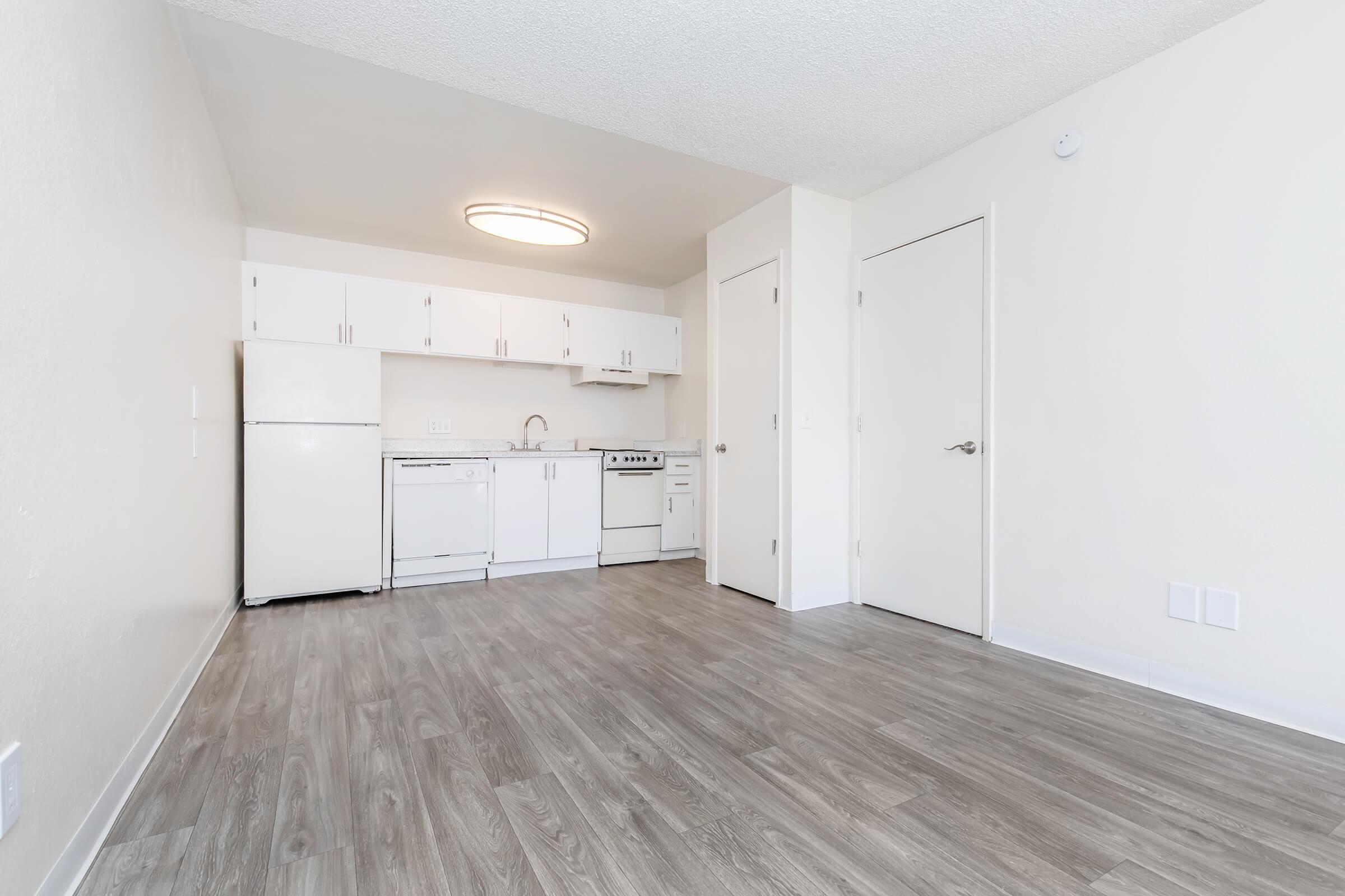 A spacious and clean kitchen area featuring white cabinets, a sink, stove, and refrigerator. The floor is made of light gray wood-like laminate. The walls are painted in a neutral tone, creating a bright and open atmosphere. A circular ceiling light provides illumination.