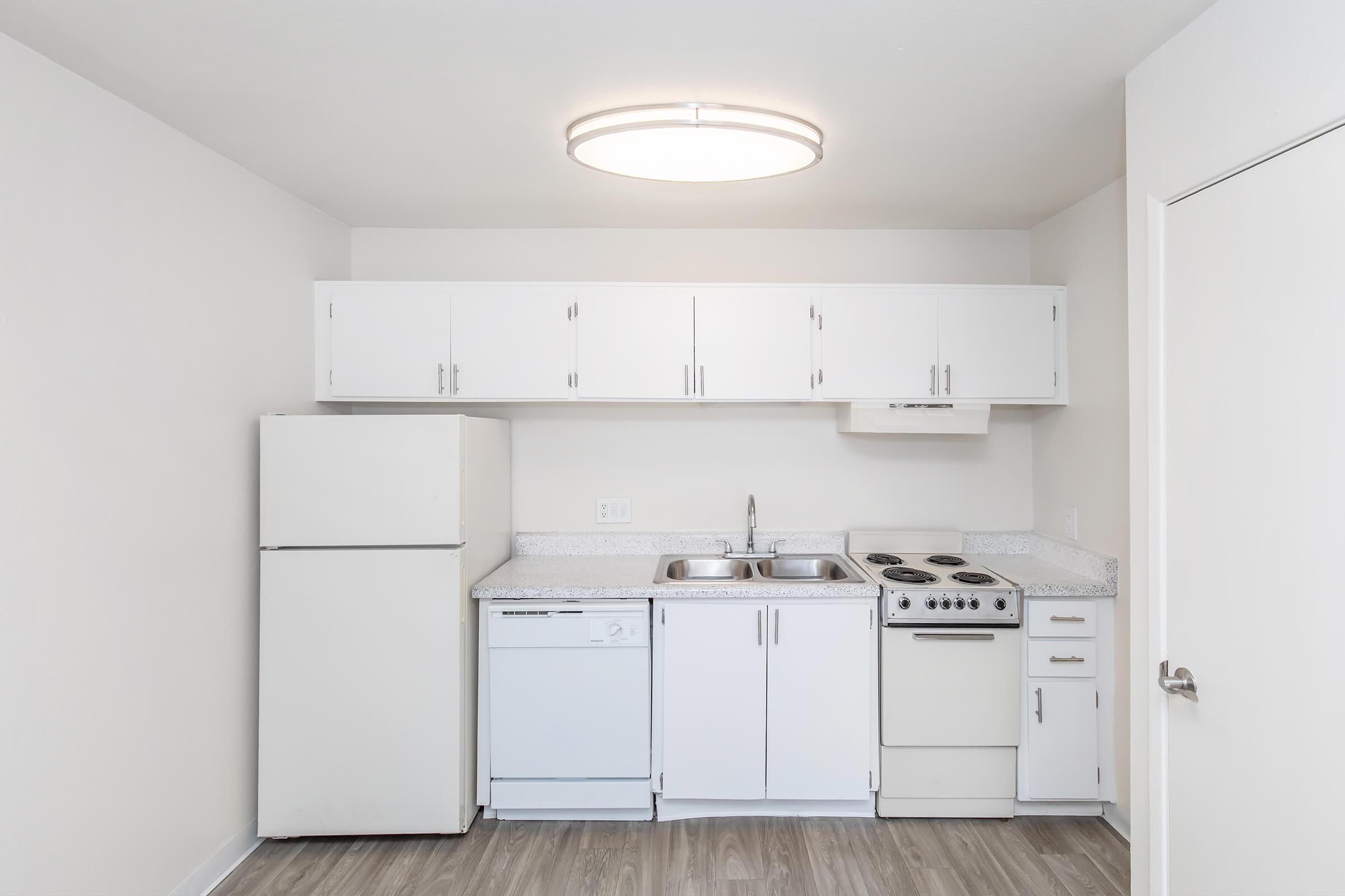A modern kitchen featuring white cabinetry, a stainless steel sink, a stove, a refrigerator, and a dishwasher. The countertop is light-colored with a textured finish. The walls are painted in a neutral shade, and a circular ceiling light provides illumination in the space. The flooring is light wood-like vinyl.