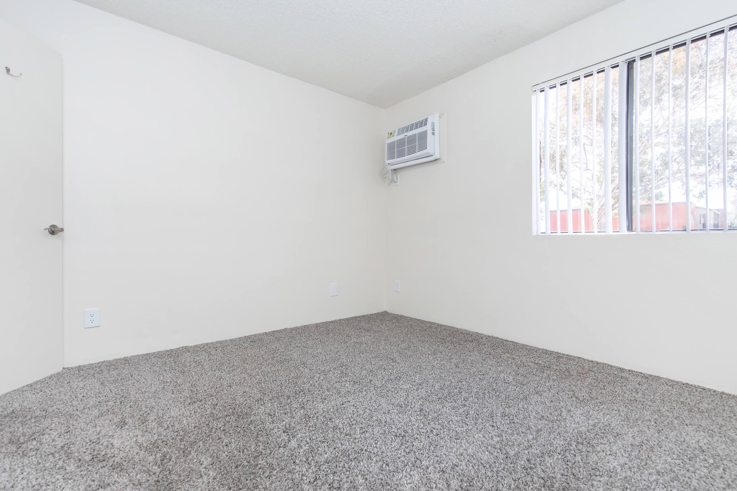 A spacious, empty room with light beige walls and a soft, carpeted floor. There is a wall-mounted air conditioning unit and a window with vertical blinds on the right side, allowing natural light to enter. A door is visible on the left side of the room, leading to an adjacent space.