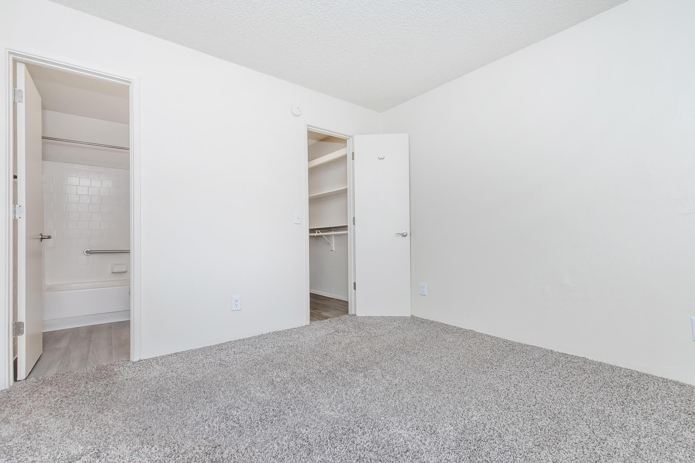 A well-lit, empty room featuring light-colored walls and carpet. There's a door on the left leading to a bathroom with a tub, and another door on the right leading to a closet with a hanging rod. The overall atmosphere is clean and spacious, ideal for personal customization.