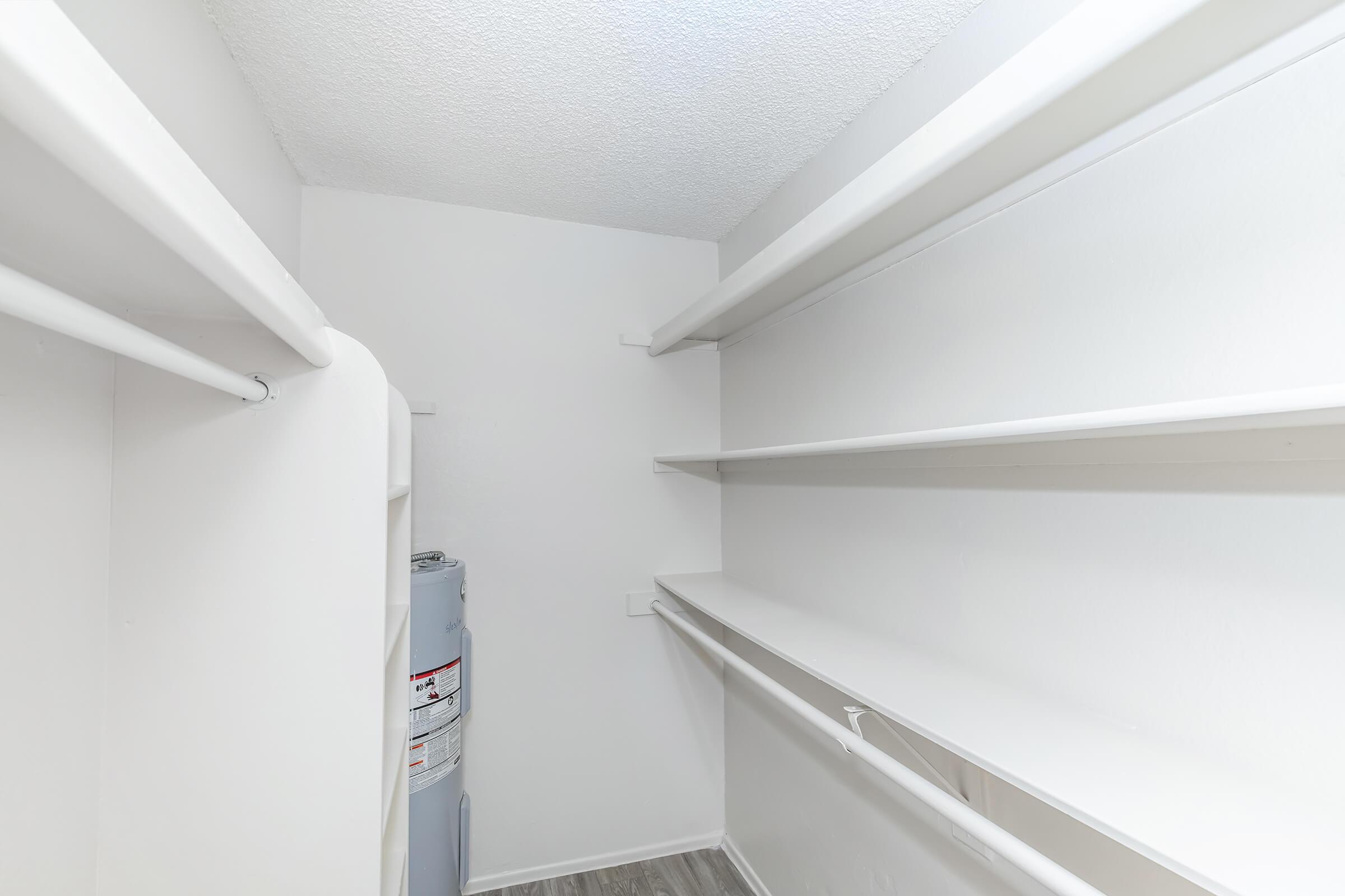 Empty closet space with white walls and shelves on either side. A hot water heater is visible in the corner. The flooring is light-colored, and the ceiling has a textured finish. The overall appearance is clean and organized, suitable for storage.