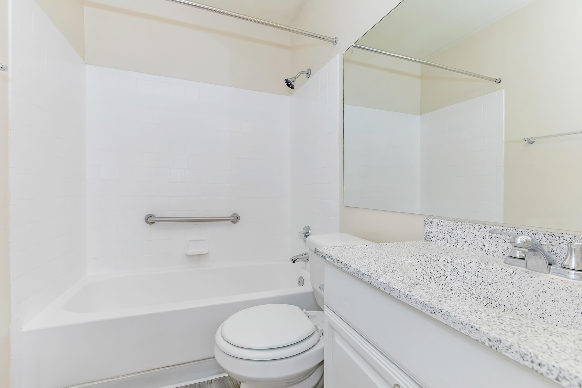 A clean and modern bathroom featuring a white tiled shower and bathtub combination, a granite countertop with a sink, a large mirror, and minimalistic fixtures. The walls are painted a light color, providing a bright and airy atmosphere. Safety grab bars are installed in the bathtub area.