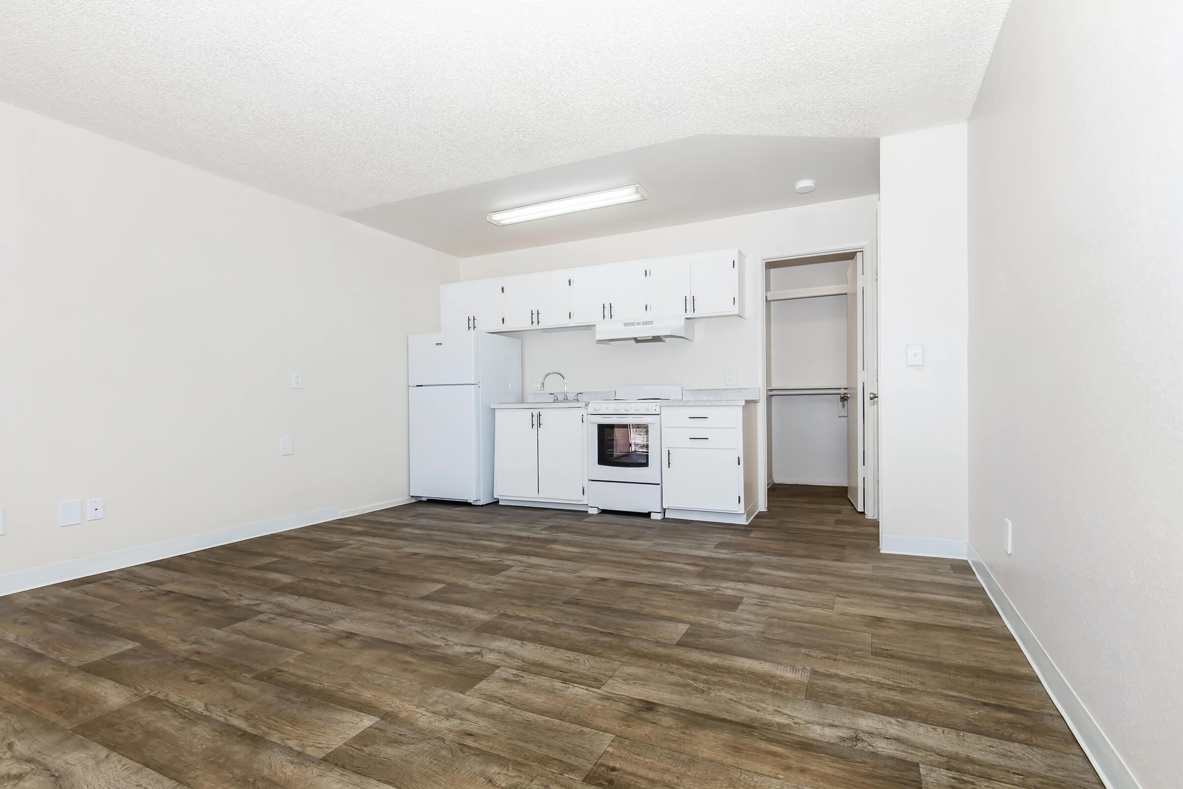 Empty interior of a small studio apartment featuring a kitchenette with white cabinets, a refrigerator, and a stove. The room has light-colored walls and a wood-style floor, creating a bright and spacious atmosphere. A closet is visible through an open door.