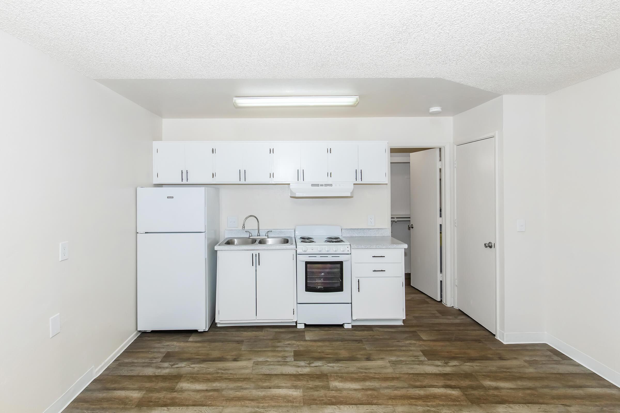 Modern kitchen area featuring white cabinetry, a refrigerator, a sink, and a stove. The walls are light-colored, and the flooring is a wood-like laminate. A doorway on the right leads to another room, and there is overhead lighting above the kitchen appliances.
