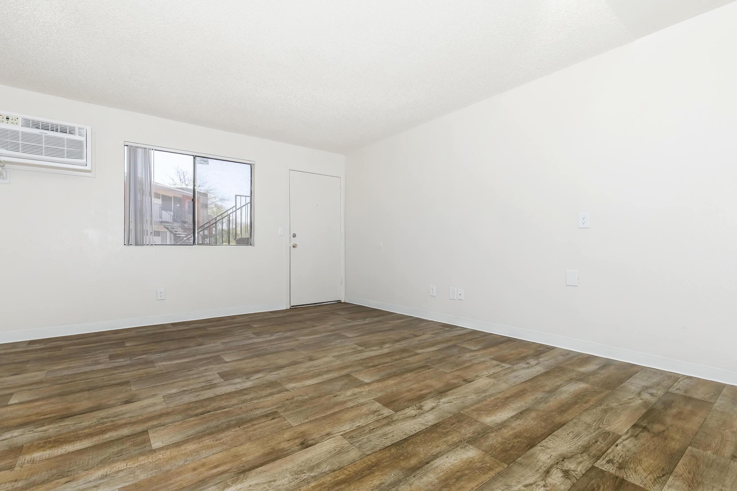 A spacious, empty room with light-colored walls and wooden flooring. A window allows natural light to enter, and a door is visible on one wall. An air conditioning unit is mounted on the wall, indicating a comfortable living space. The room is clean and well-maintained, suitable for various furnishings.