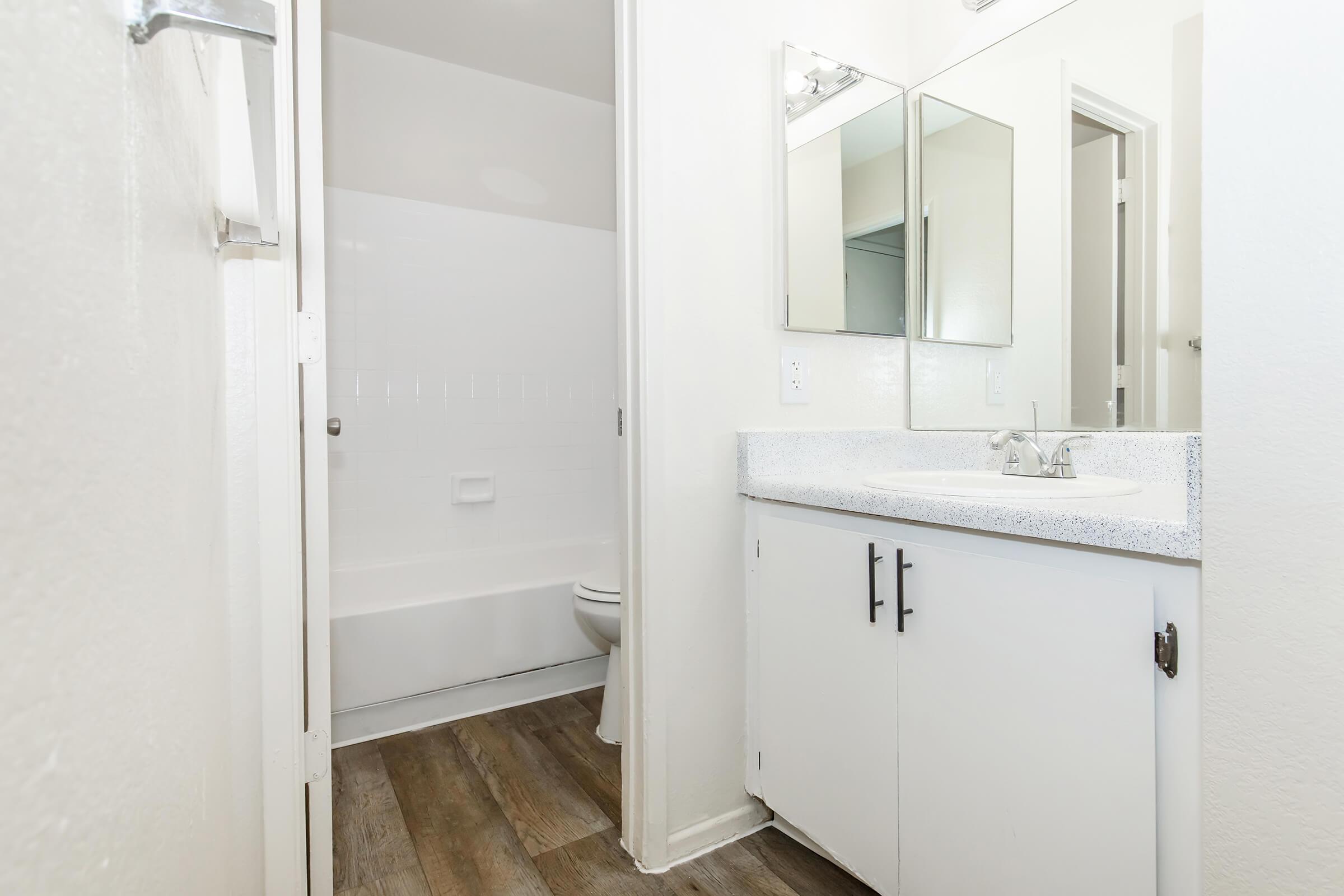 A clean and modern bathroom featuring a white bathtub and shower combination, a double vanity with sinks and mirrors above, light-colored walls, and laminate flooring. The space is well-lit and has a minimalist design with two mirrors and a door leading to another room.