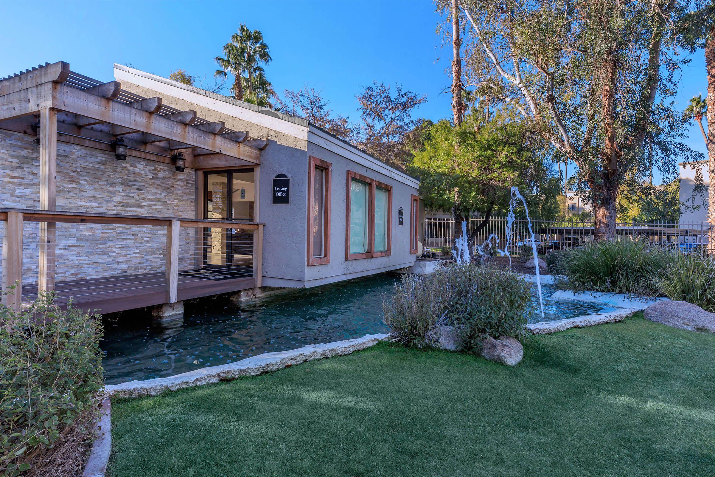 A modern building with a stone facade, surrounded by a landscaped area featuring a water fountain, grass, and trees. The structure is adjacent to a calm body of water, with large windows reflecting the clear blue sky.