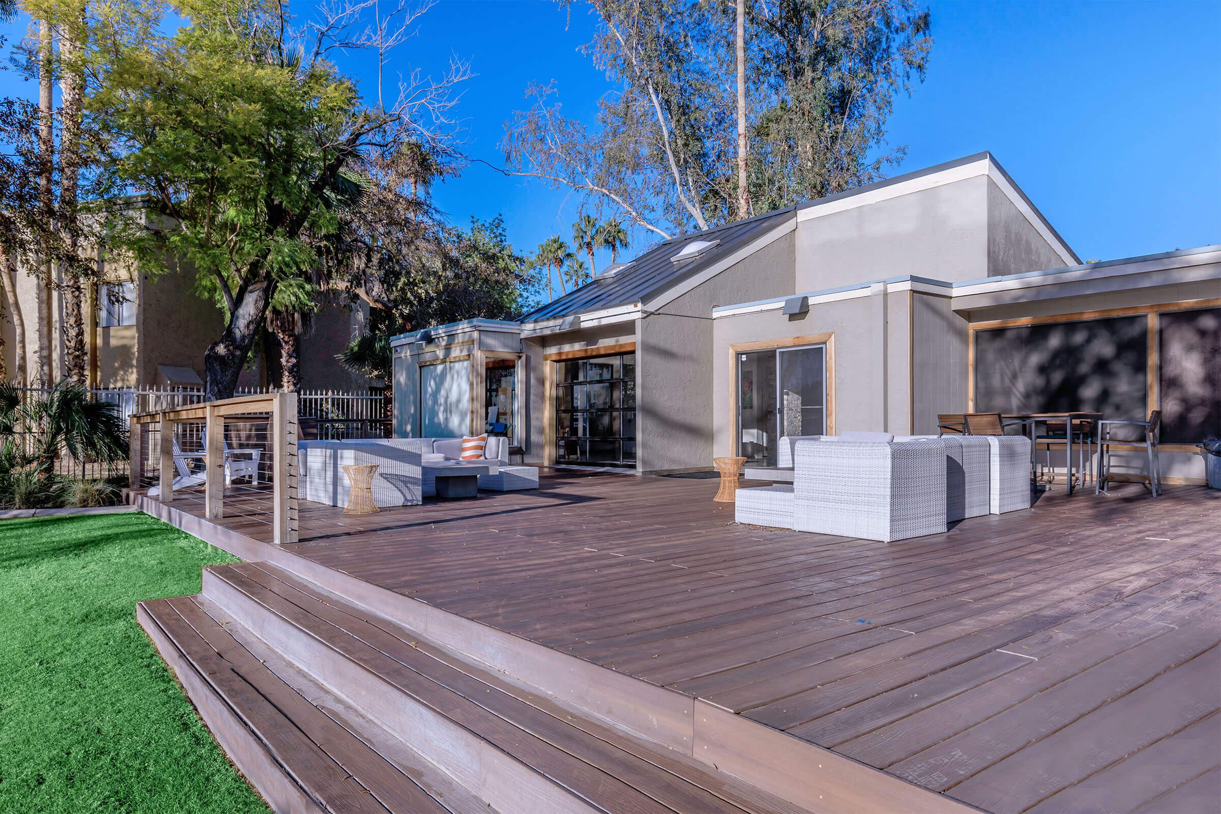 A modern outdoor patio area featuring wooden decking, comfortable seating arrangements, and greenery. The space is designed for relaxation and social gatherings, with clear blue skies in the background.