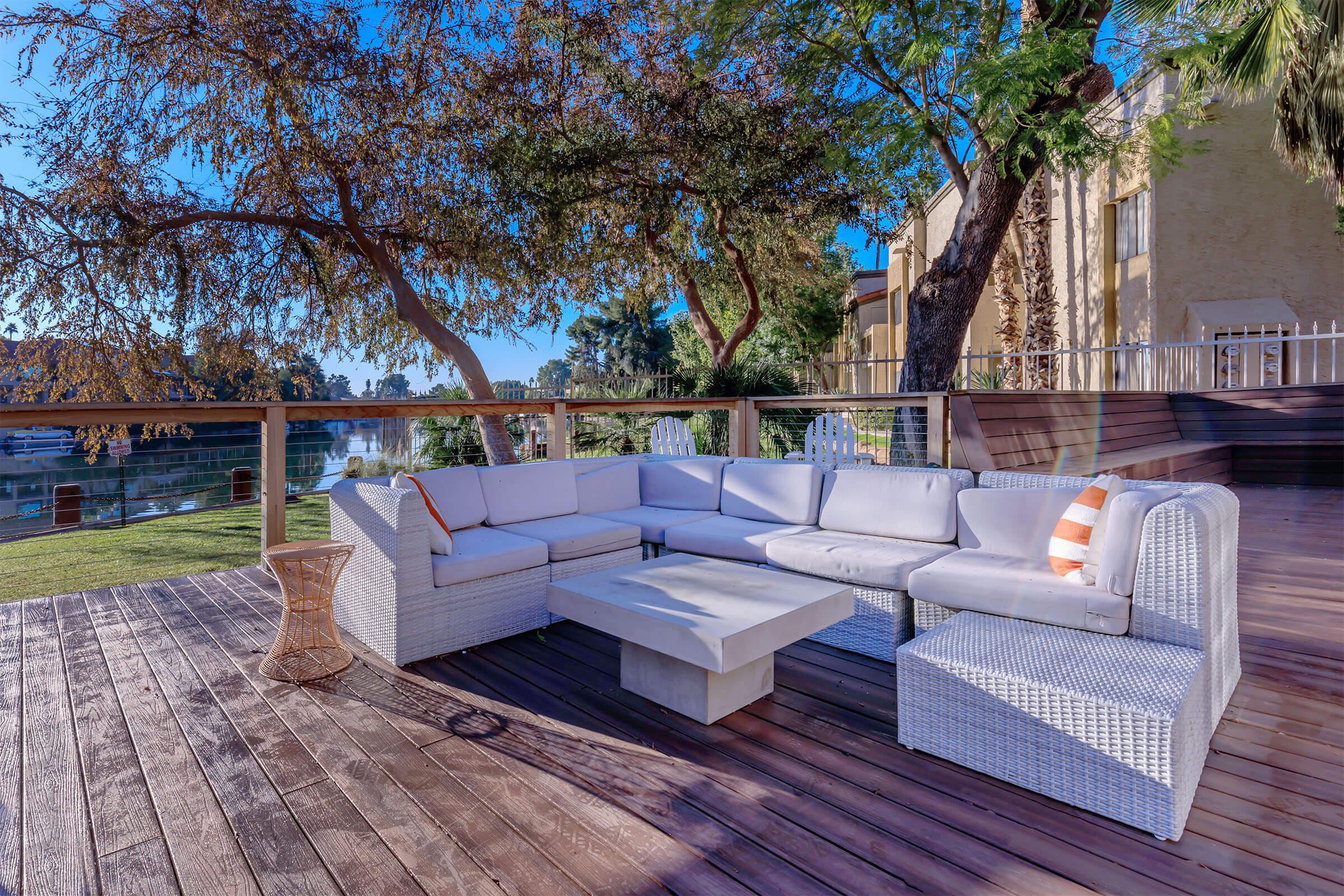 A spacious outdoor seating area featuring a white sectional sofa with orange accents, a light gray coffee table, and a small side table, set on a wooden deck surrounded by trees and a serene body of water in the background.