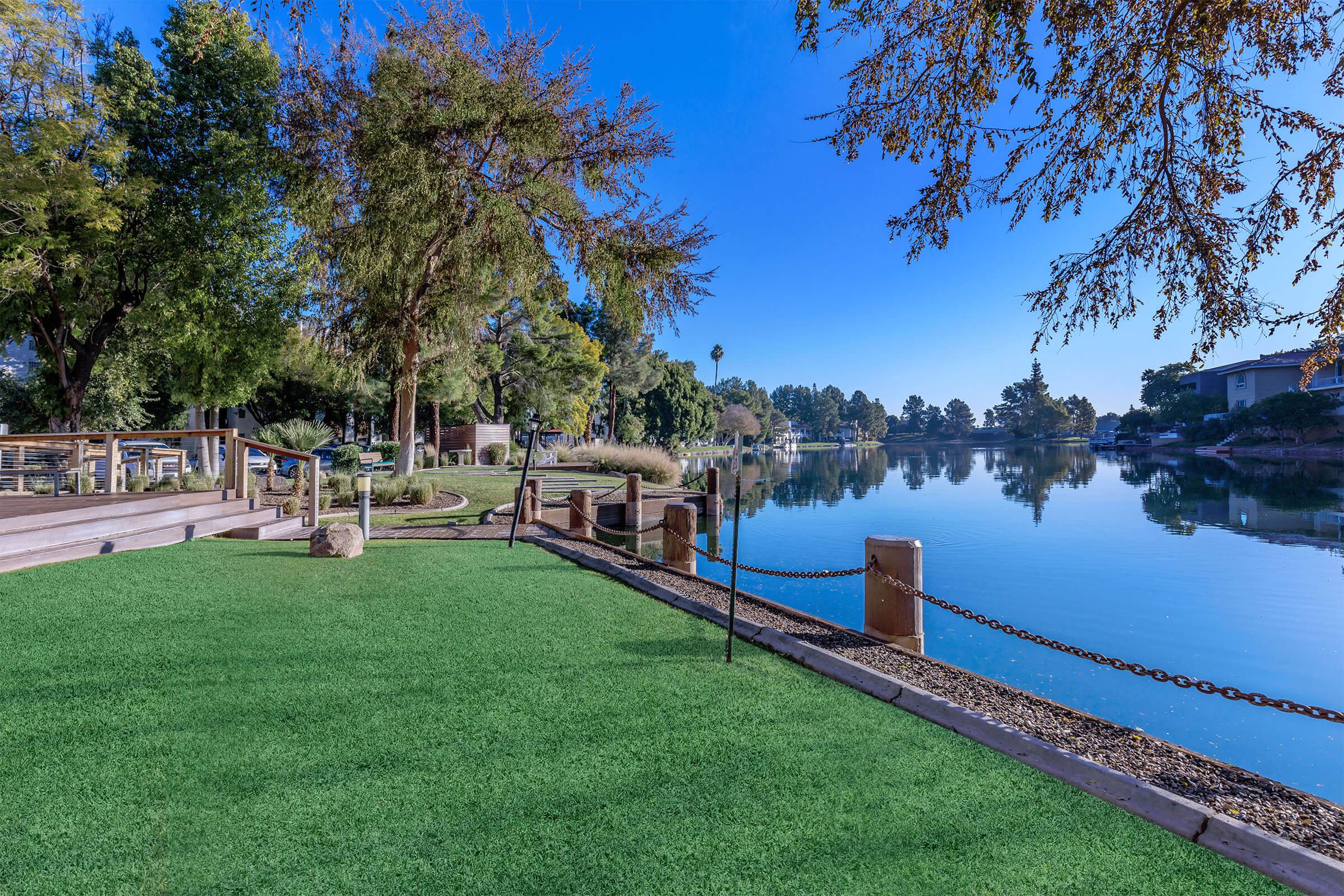 A tranquil lakeside scene featuring lush green grass, trees, and a clear blue sky, with calm water reflecting the surroundings. There are pathways and posts along the lake, creating a serene outdoor space.