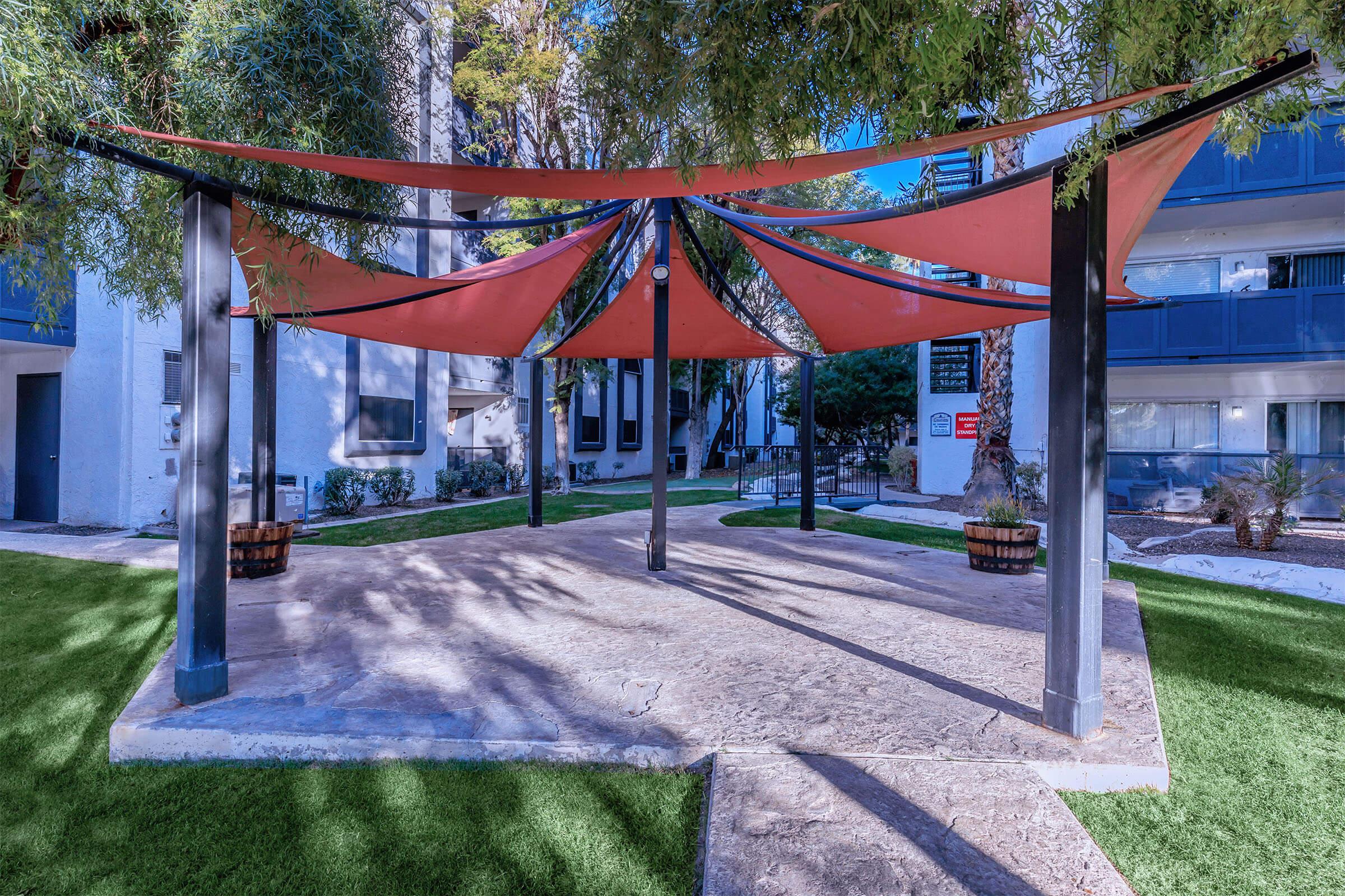 Outdoor shaded area featuring a canopy with red fabric stretched over a stone patio, surrounded by green grass and shrubbery. Modern apartment buildings are visible in the background.