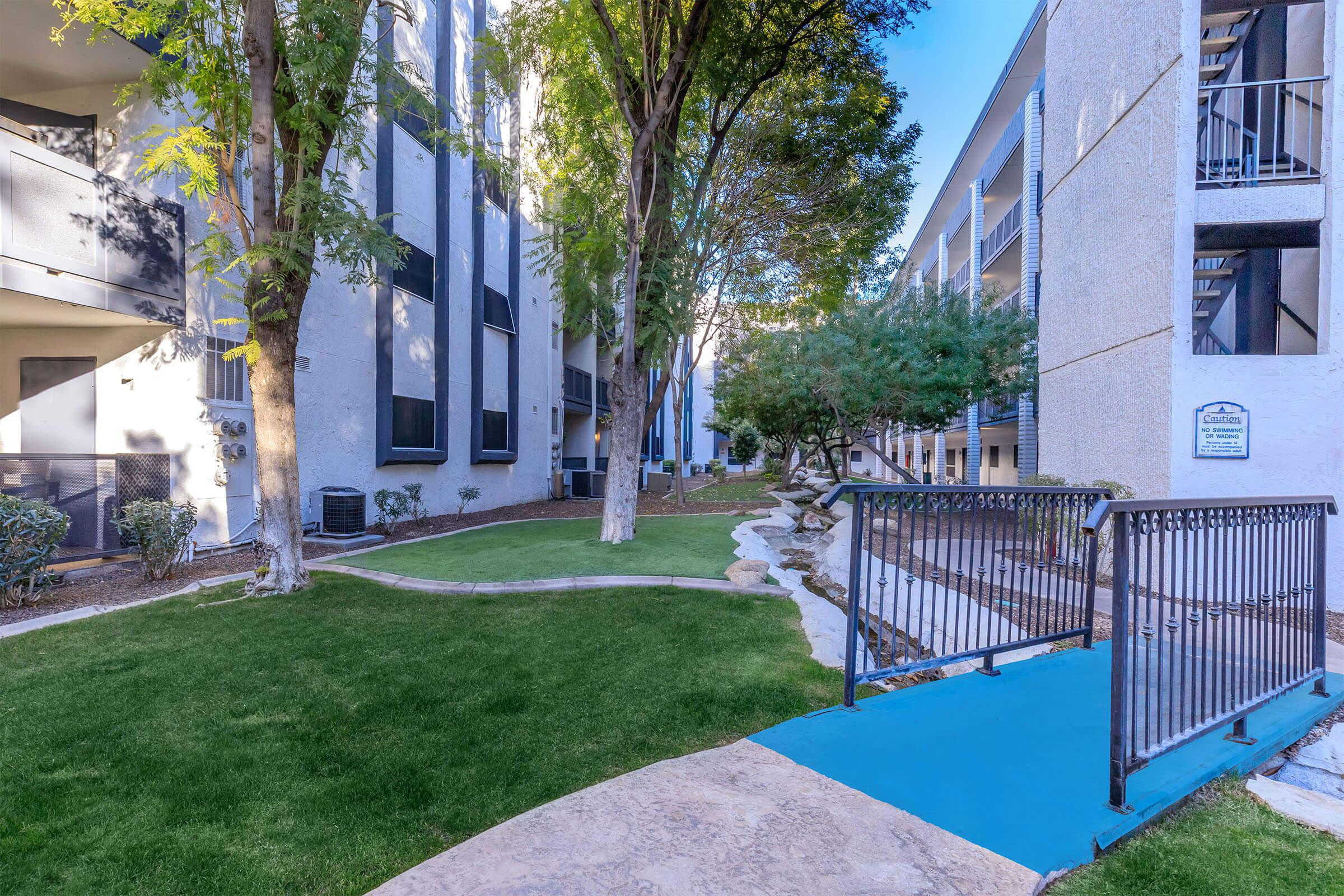 A landscaped area featuring green grass, trees, and a small blue bridge, with modern apartment buildings in the background. The scene is well-lit, suggesting a sunny day.